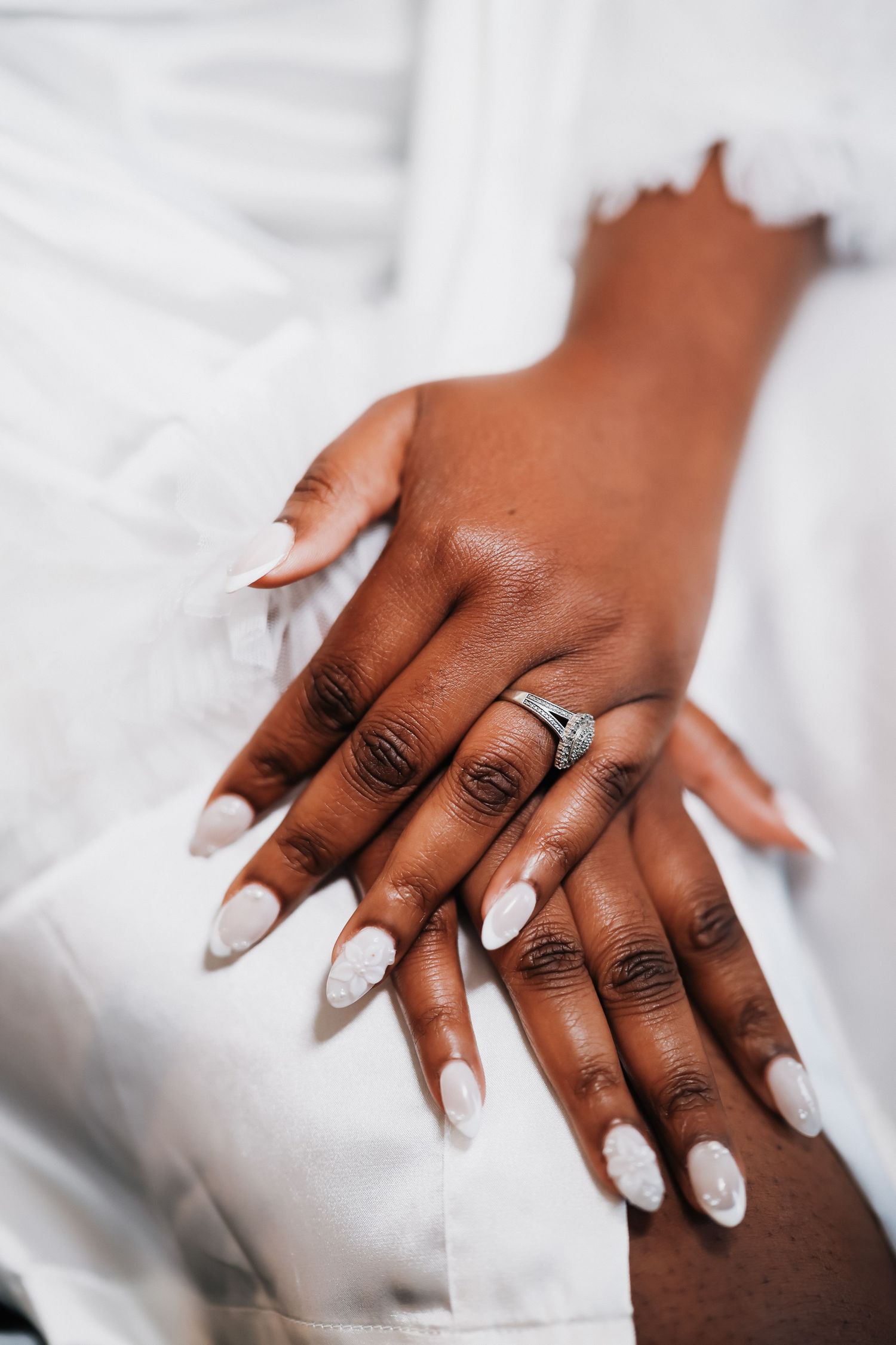 Close-up of a bride's hands with white almond nails and a diamond engagement ring, resting on a white dress.
