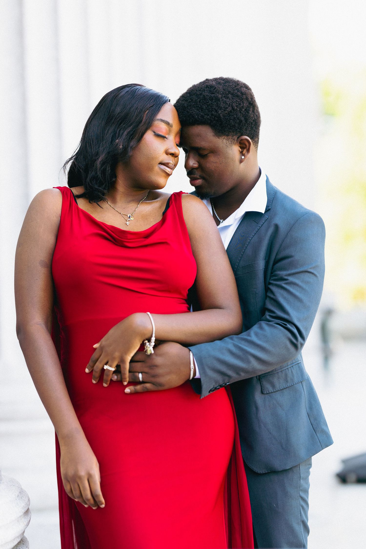 A couple in formal wear embraces tenderly, woman in red gown, man in gray suit, sharing an intimate moment outdoors.
