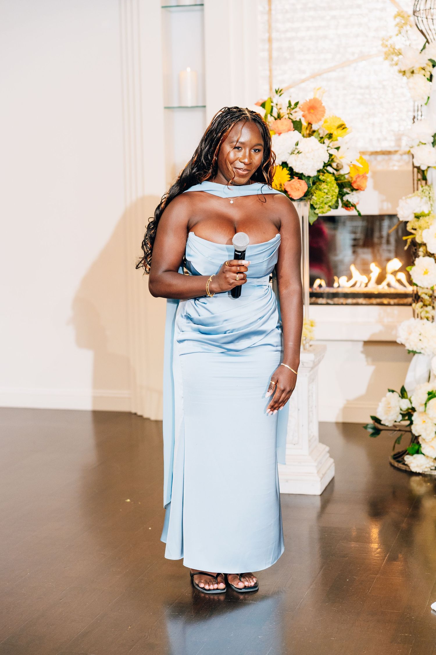 Woman in elegant light blue strapless gown holding microphone while giving a speech at a wedding reception.
