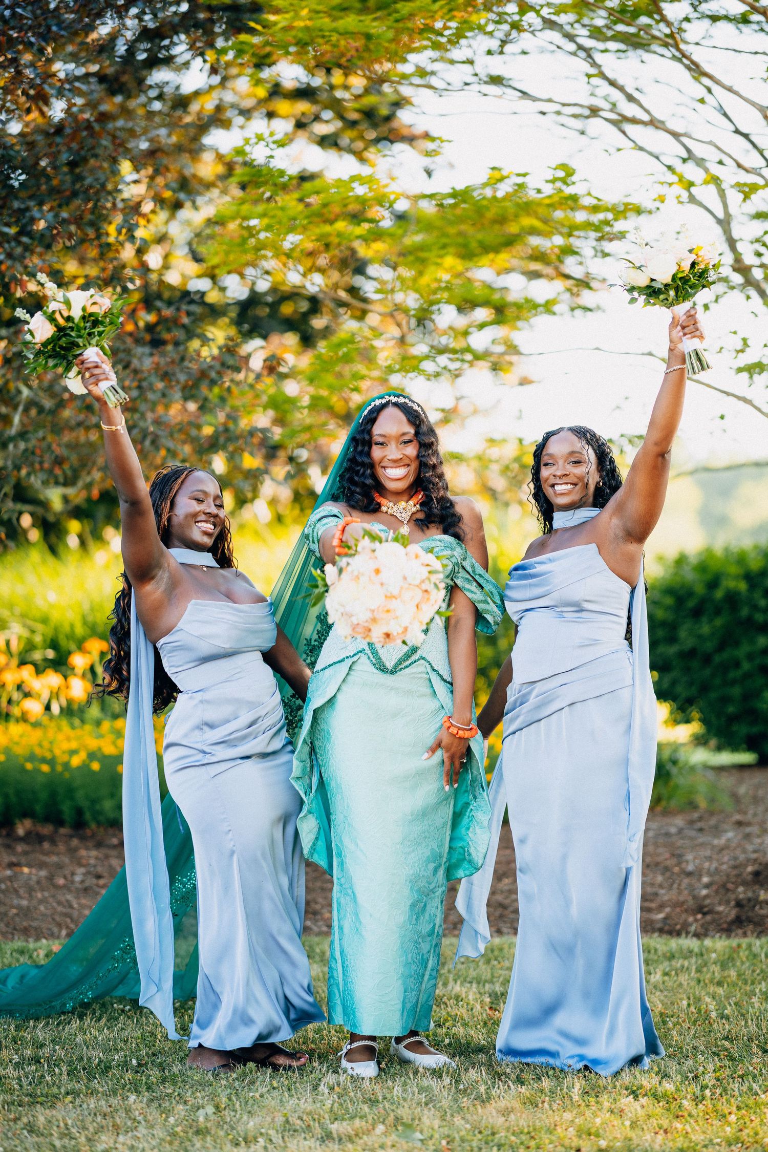 Bride in teal gown with two bridesmaids in blue dresses celebrating outdoors, holding bouquets.
