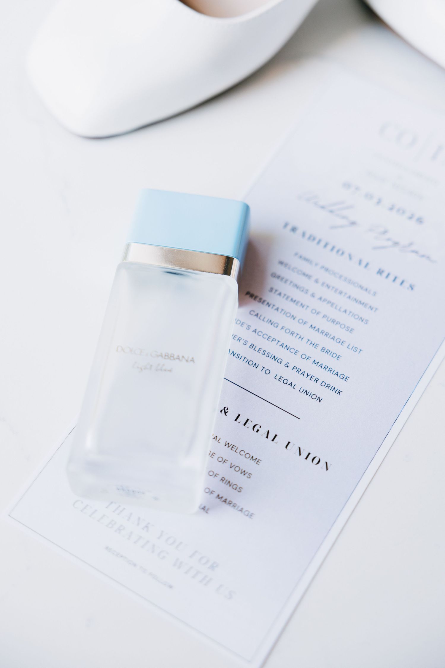 Dolce & Gabbana Light Blue perfume bottle resting on a wedding ceremony program beside white bridal heels.