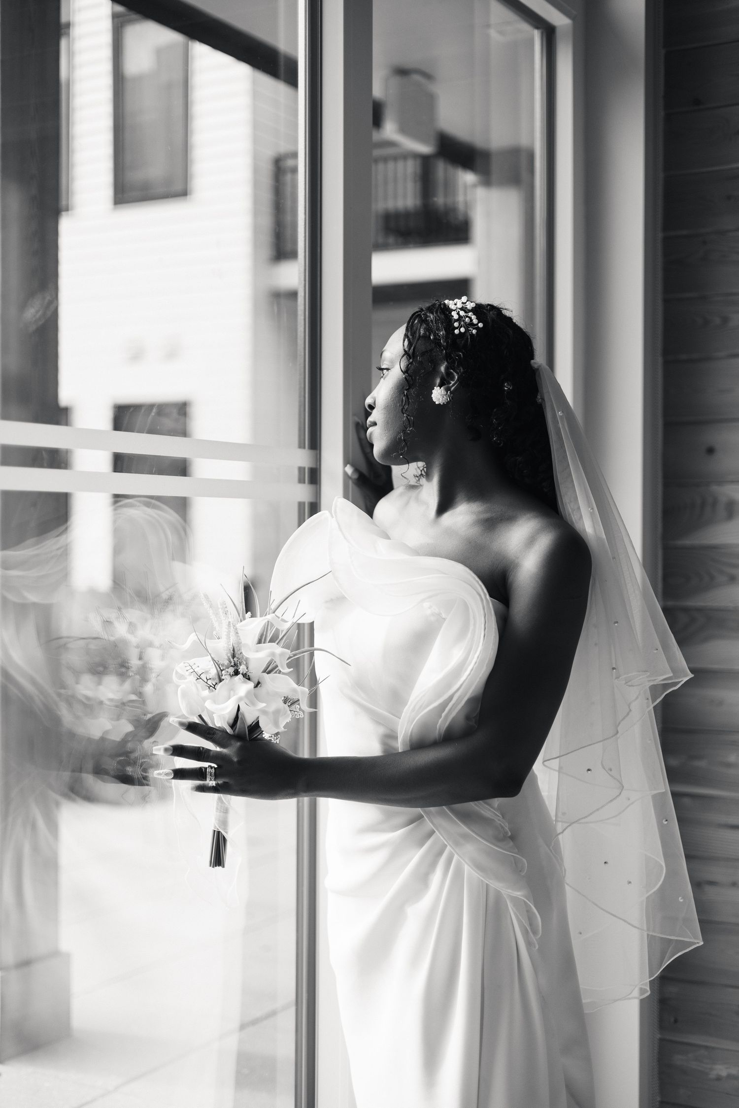 Elegant Black bride in white gown and veil holds bouquet near window in black and white photo.