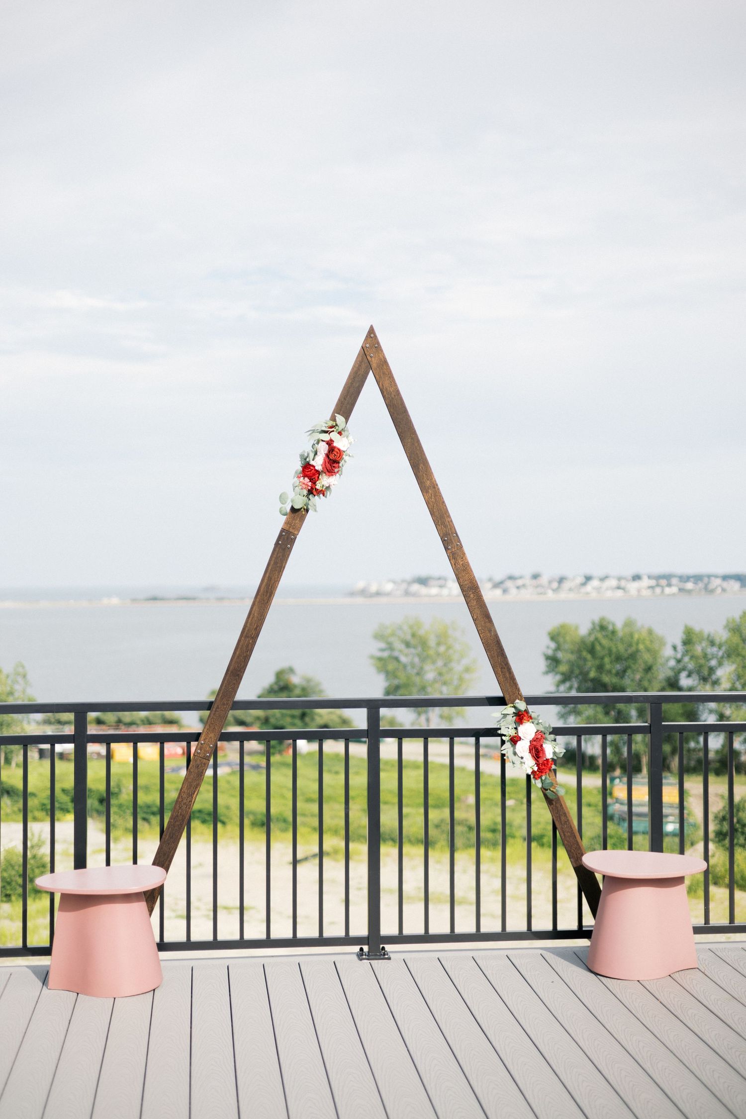 Triangular wooden wedding arch with floral accents on rooftop deck overlooking waterfront, flanked by pink pedestals.