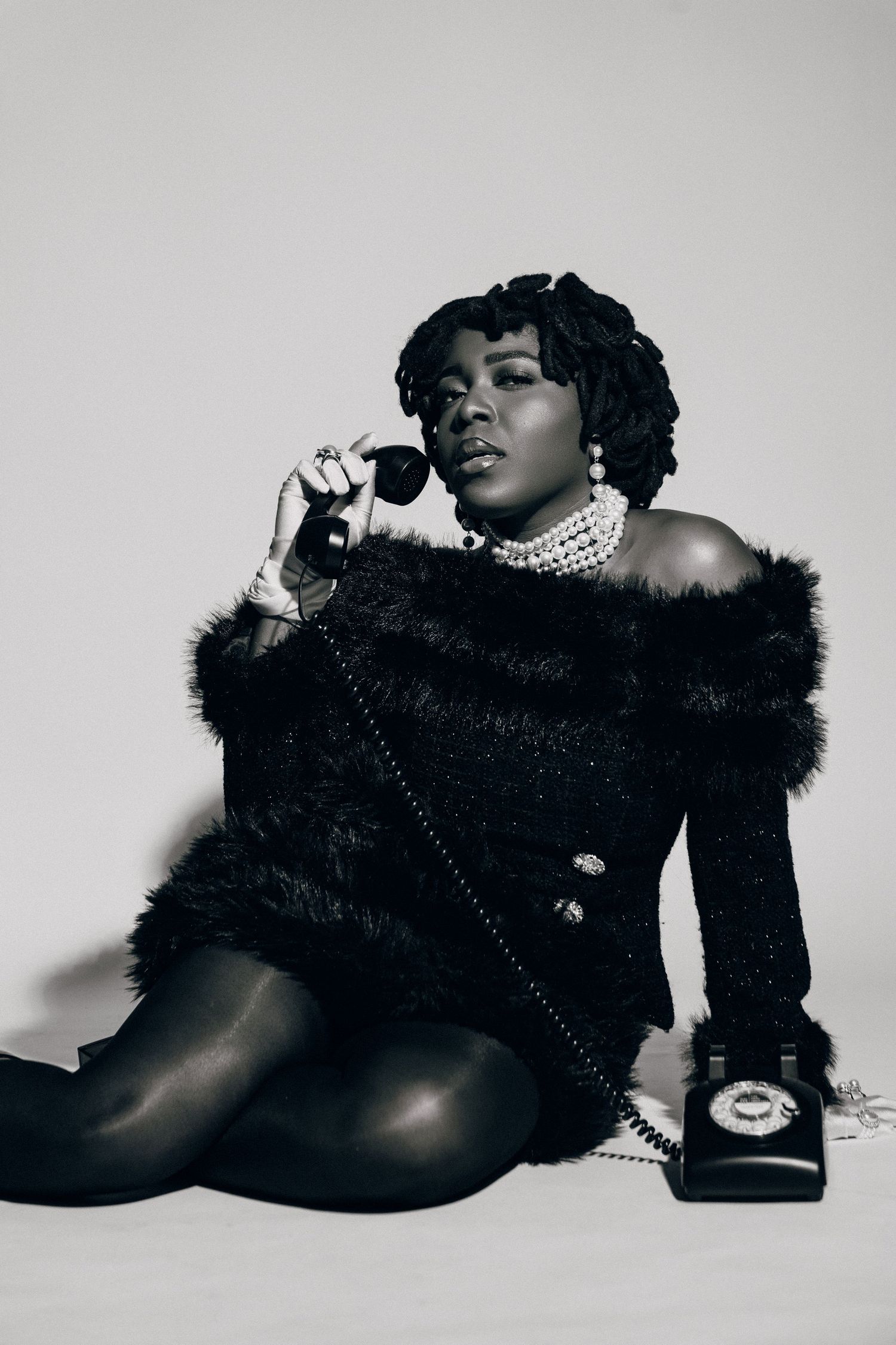 Black and white photo of a woman in fur off-shoulder top holding vintage rotary phone receiver, seated on floor.