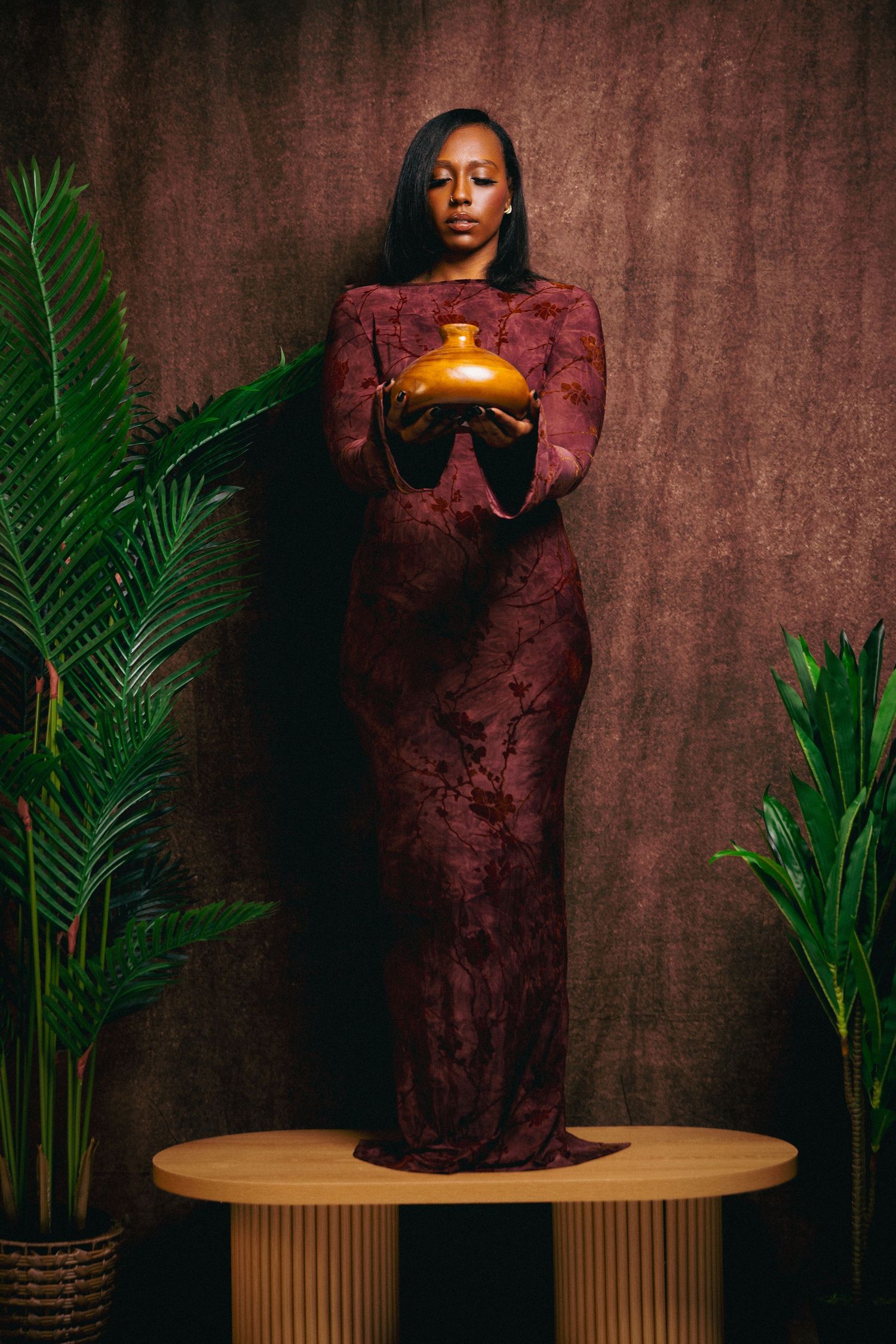 A Black woman in a burgundy lace gown holds a golden vase while standing on a wooden pedestal surrounded by tropical plants.