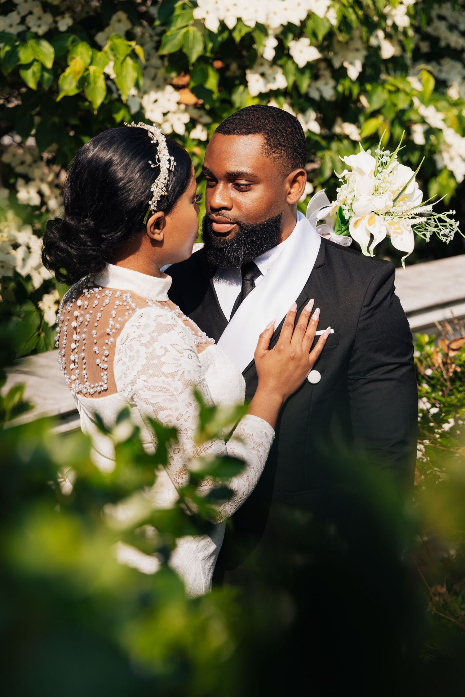 Elegant Black couple in wedding attire embrace outdoors, bride in pearl gown, groom in black tuxedo with white bouquet.