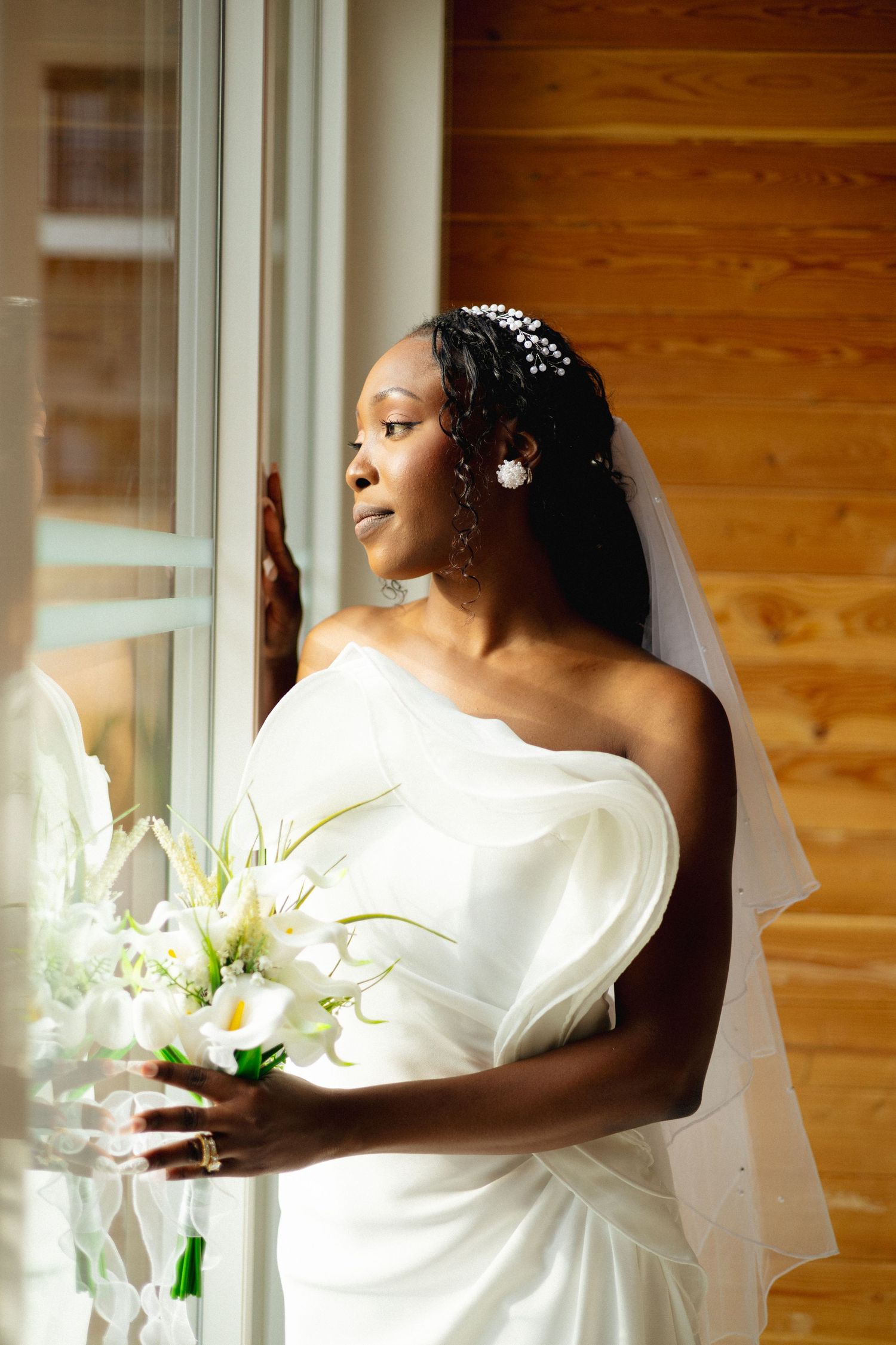 Beautiful Black bride in white one-shoulder wedding dress holding calla lily bouquet beside sunlit window.