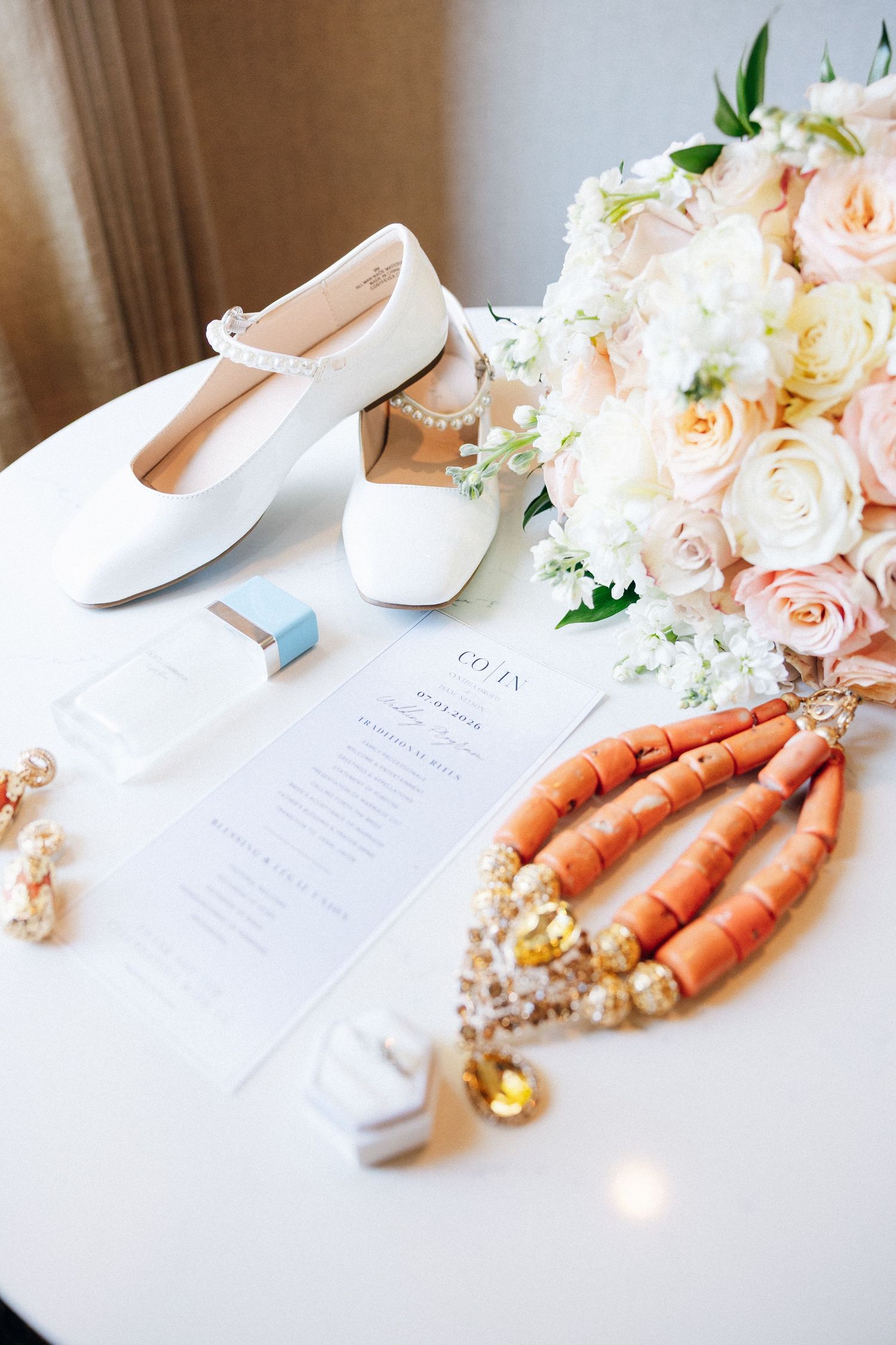 Wedding flat lay with white bridal shoes, coral beaded necklace, bouquet, and ceremony program on table.