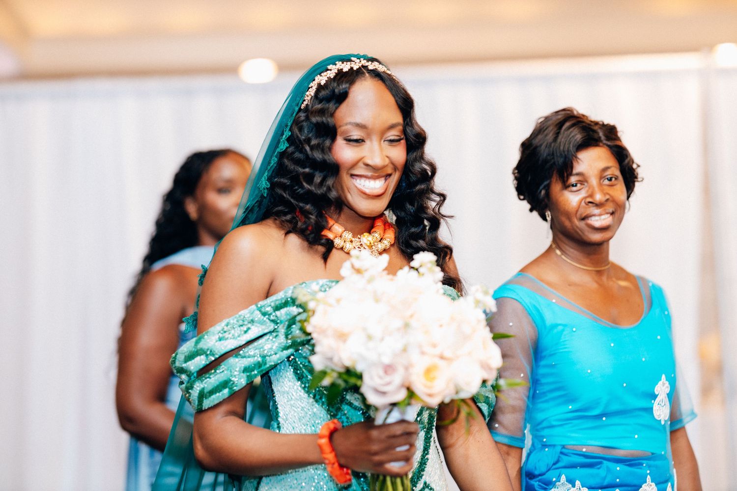A joyful bride in a teal African wedding dress and veil walks down the aisle holding white flowers.