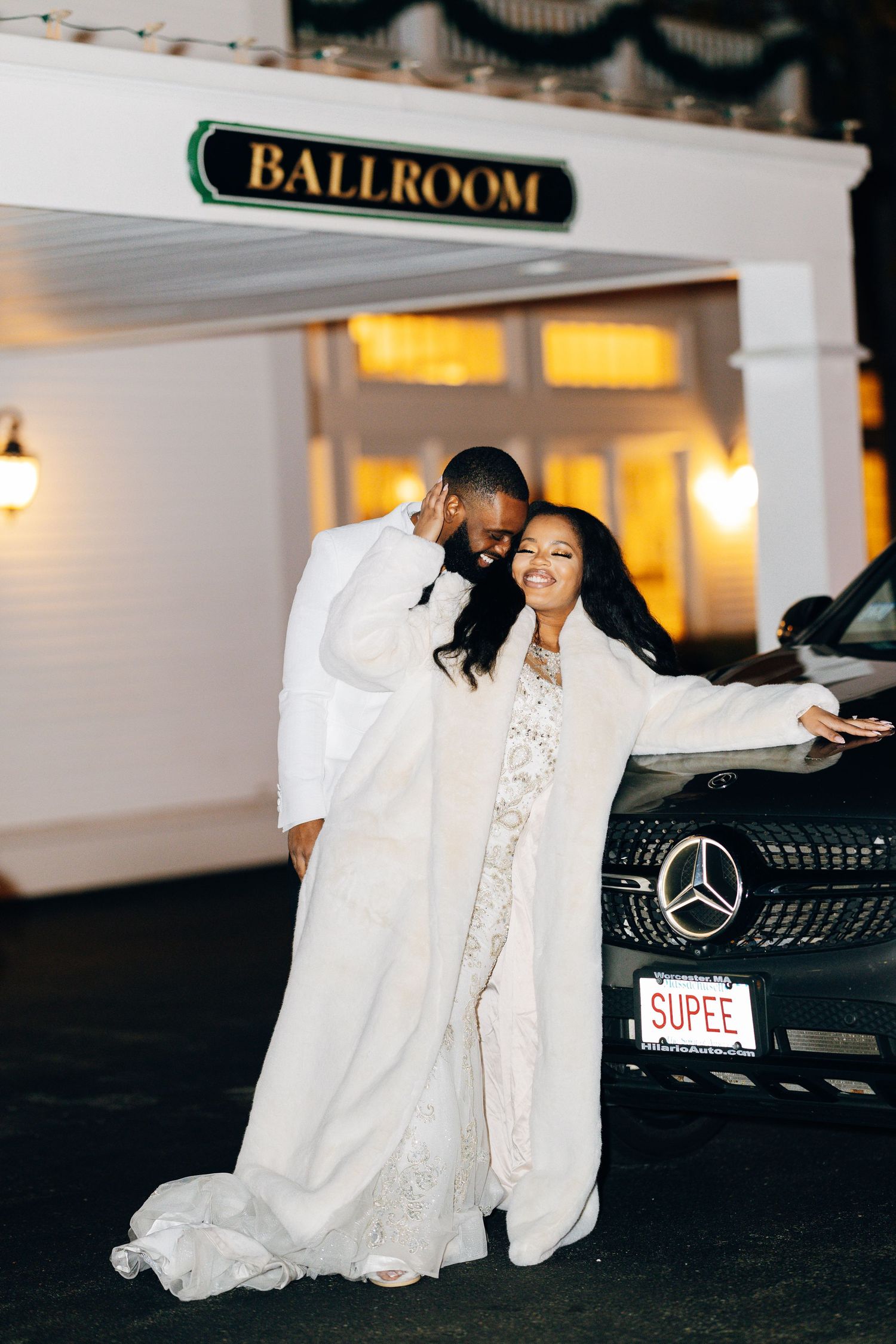 Newlyweds pose together in white wedding attire outside a ballroom entrance beside a black Mercedes.