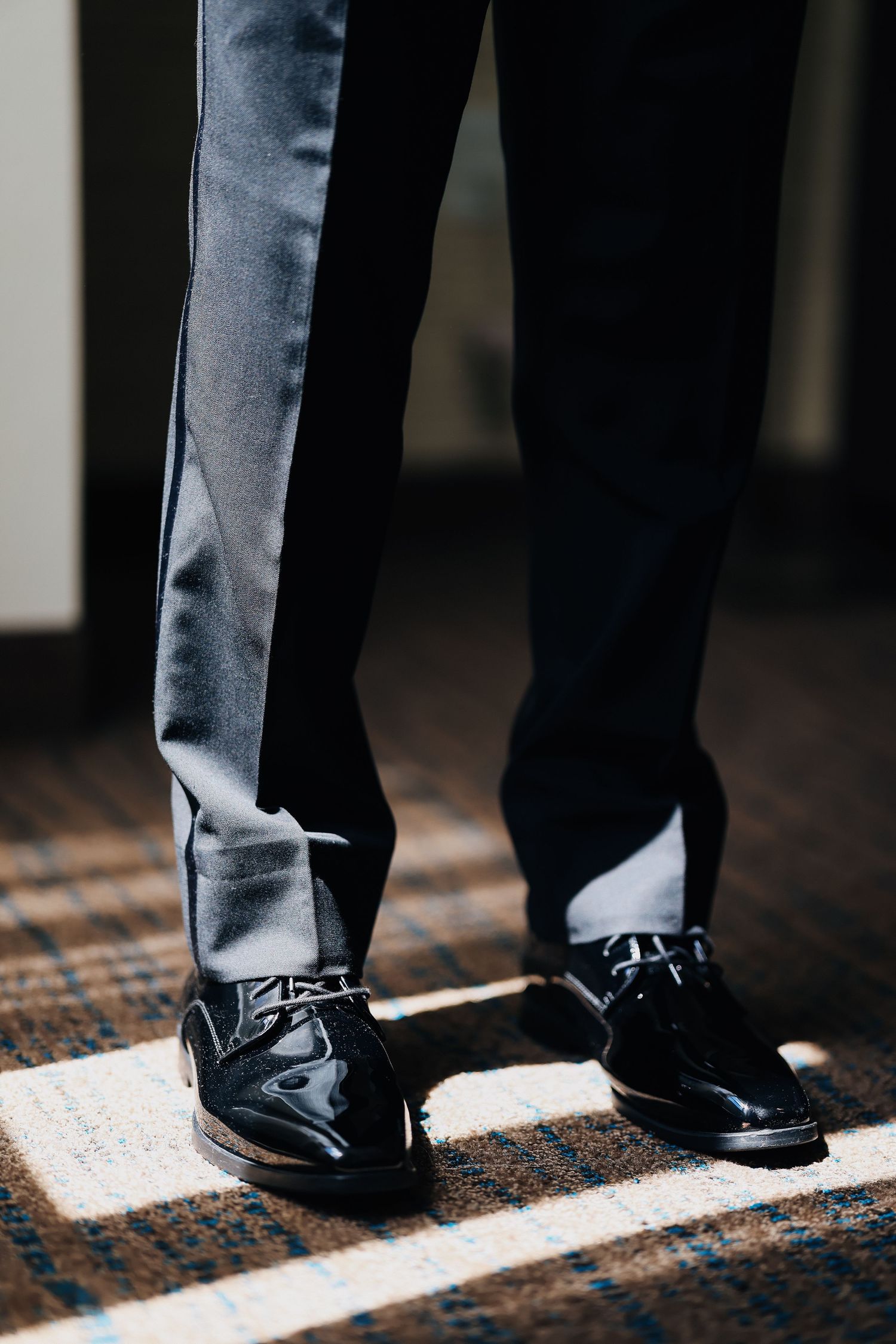 Close-up of a groom wearing black patent leather dress shoes and dark tuxedo pants standing on a patterned carpet.