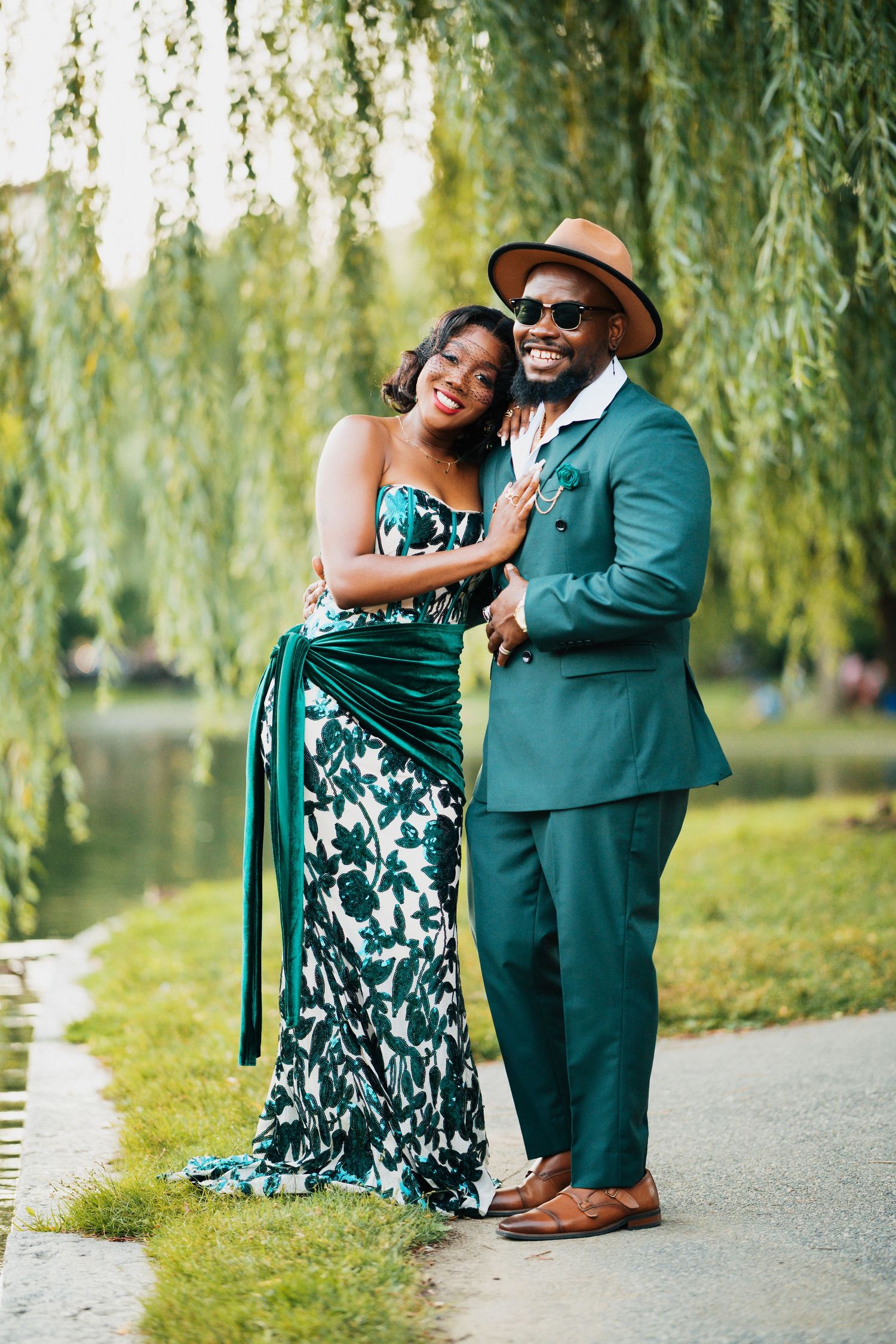 Smiling Black couple in matching green outfits pose by a willow tree near a lake.