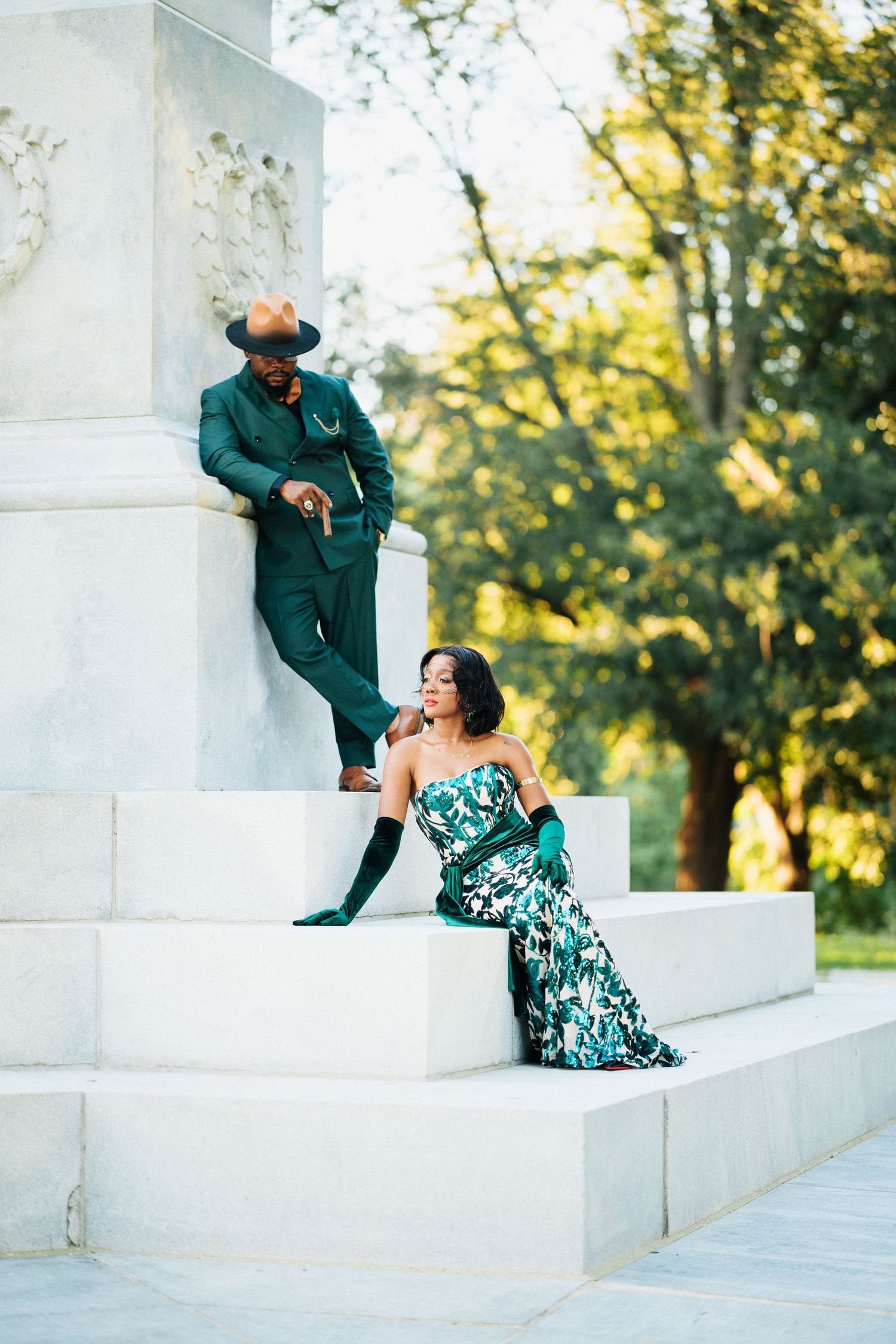 Stylish couple in coordinating emerald green outfits posing on white marble steps in a sunlit park setting.