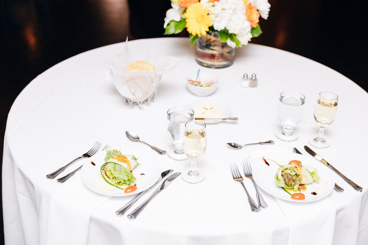 Elegant white round table set for two with salad plates, silverware, wine glasses, and a floral centerpiece.