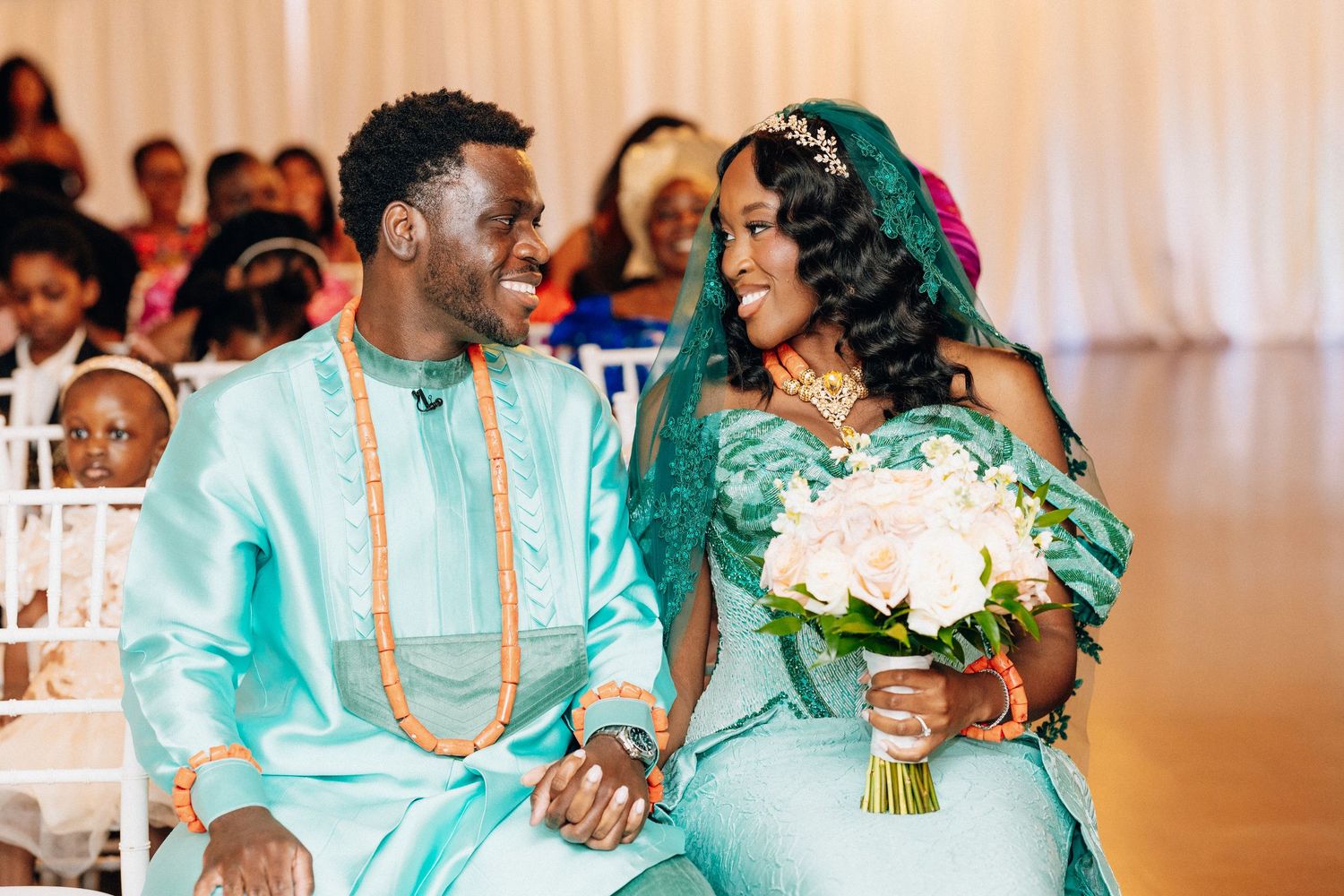 A bride and groom in matching teal African wedding attire sit together holding hands at ceremony.