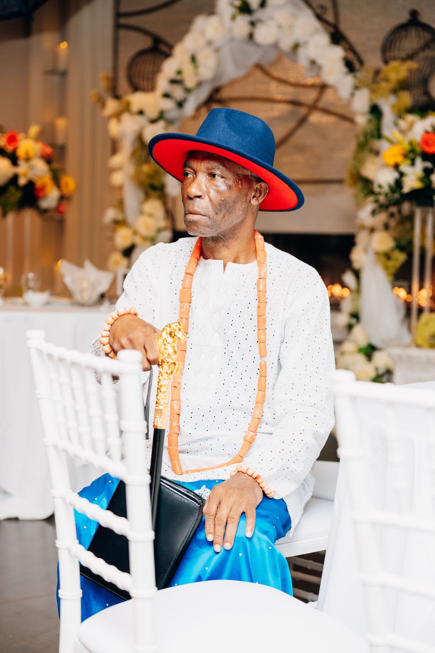 An elderly African man in traditional attire with blue and red hat, orange beaded necklace, seated at a decorated event venue.