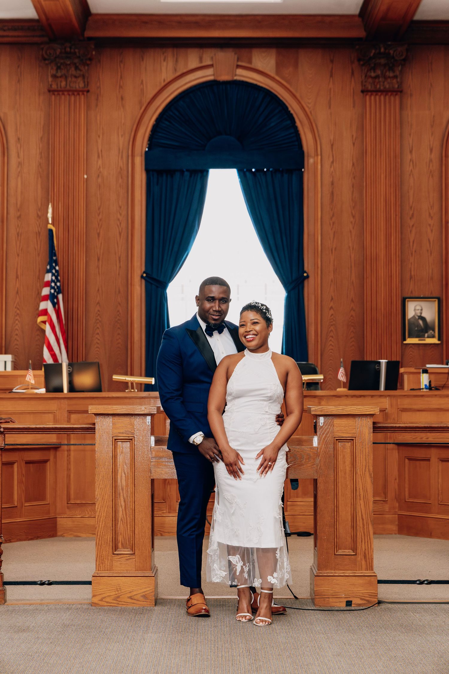 Couple in wedding attire posing in elegant wood-paneled courthouse with American flag.