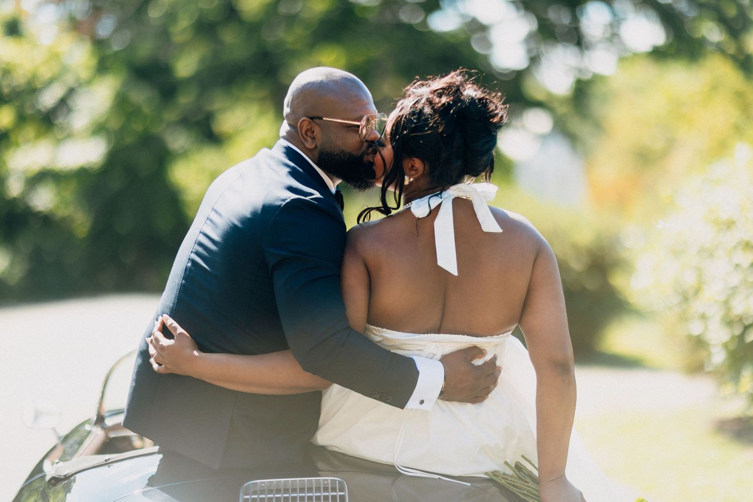 Groom in navy suit shares a romantic kiss with bride in white halter wedding dress outdoors near vintage car.