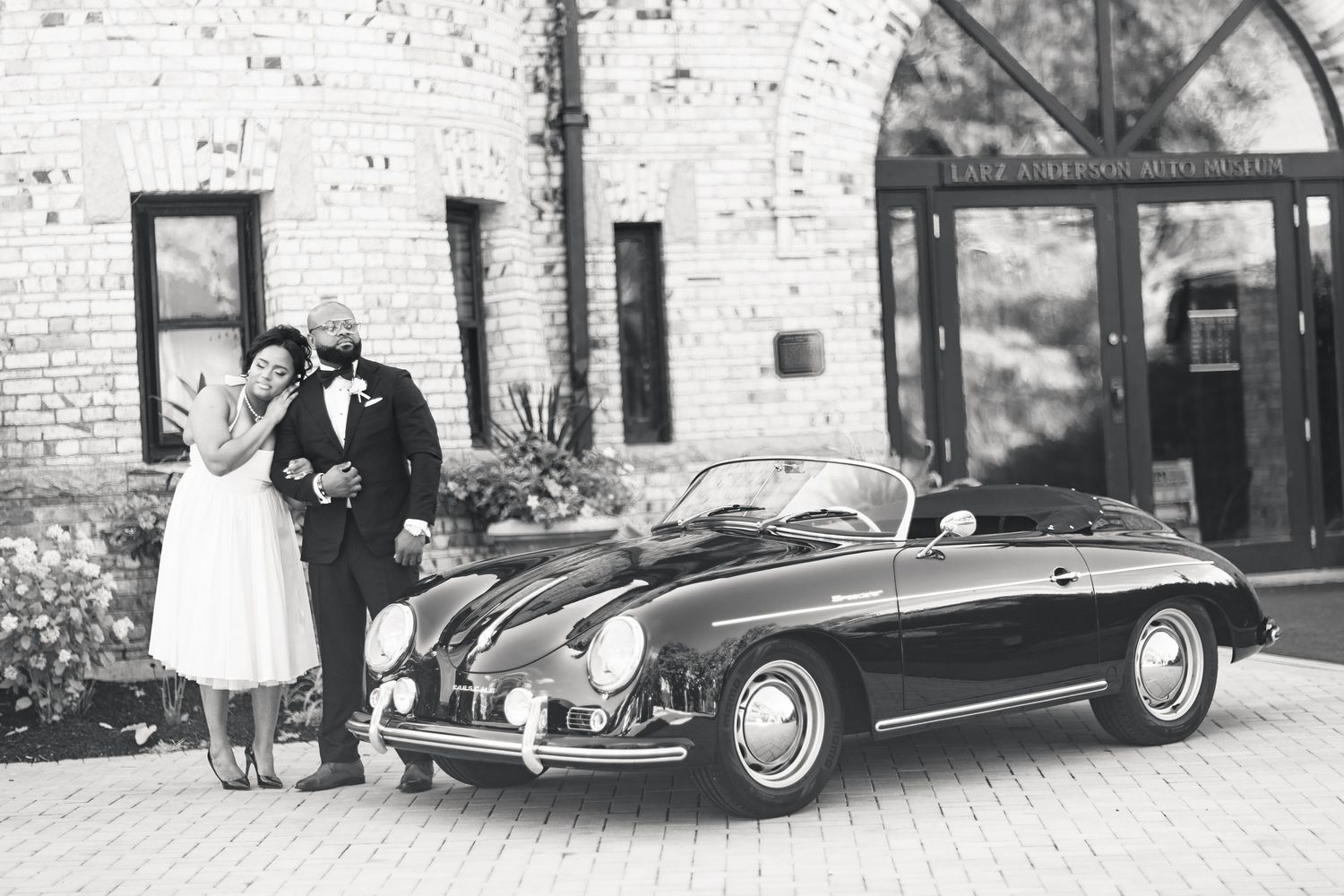 Black and white photo of newlyweds posing with a classic vintage Porsche 356 convertible.