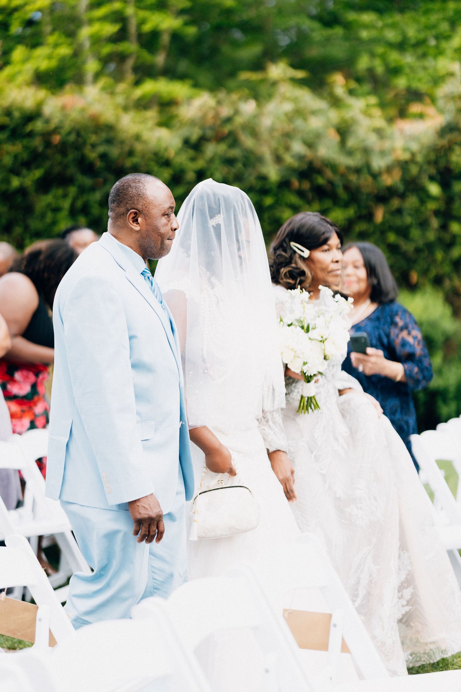 A bride in a white lace gown and veil walks down the aisle with a man in a light blue suit at an outdoor wedding.