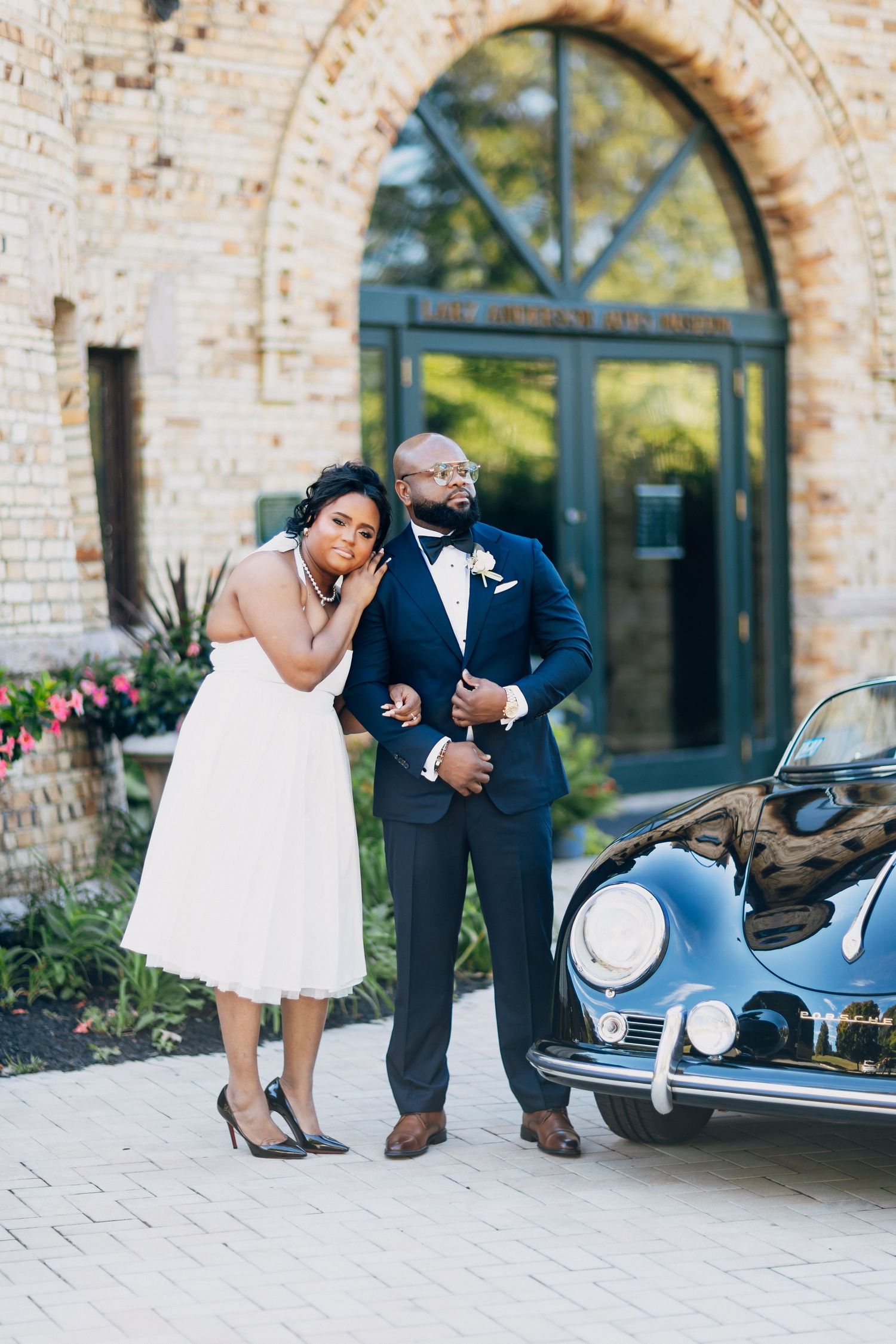 African American bride and groom posing elegantly beside a vintage black Porsche outside a brick building.