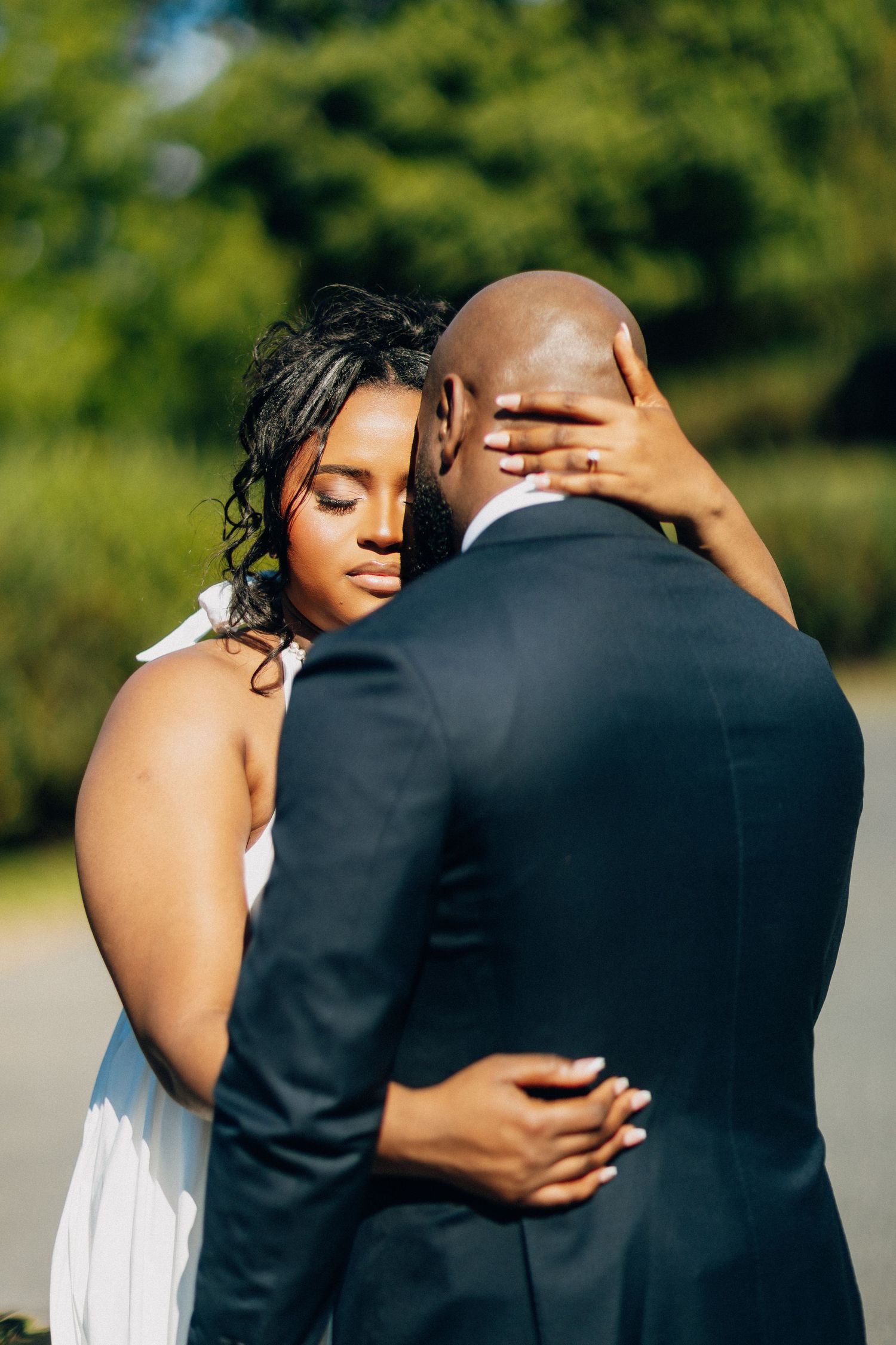 Bride in white halter gown embracing groom in navy suit from behind in a romantic outdoor wedding portrait.