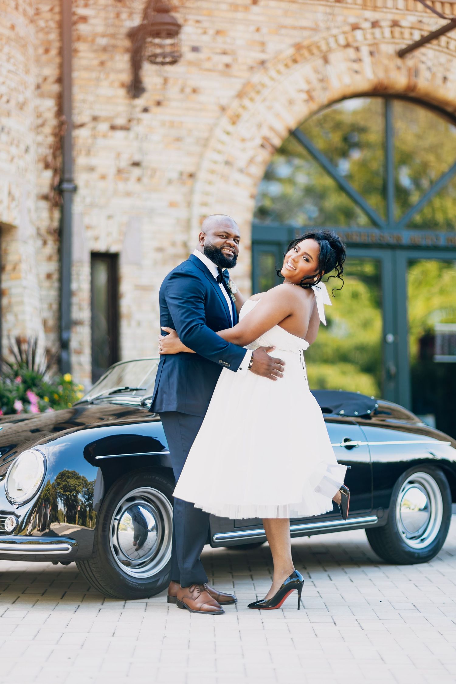 Smiling African American bride and groom hugging beside a vintage black Porsche at their wedding.