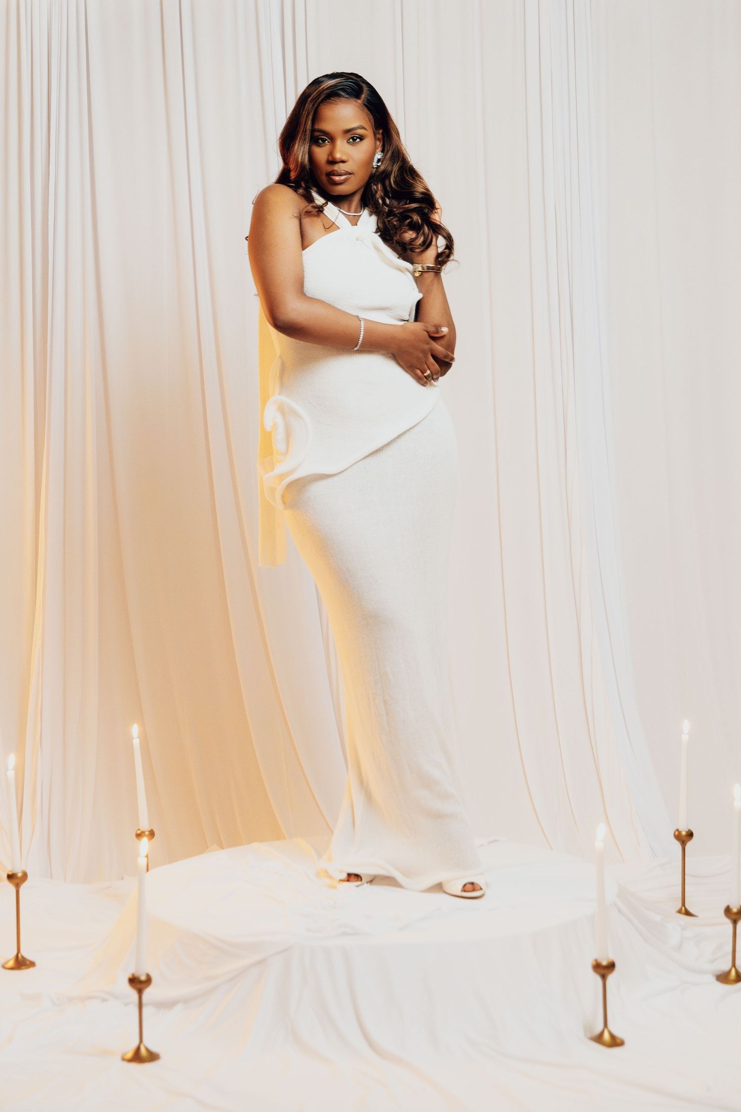 Elegant woman in white halter jumpsuit poses on circular platform with candles and draped fabric backdrop.