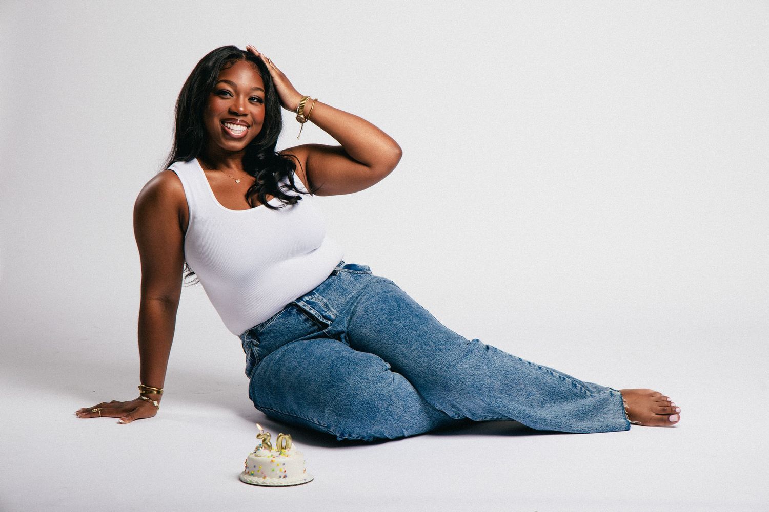 Woman in white tank top and blue jeans sitting on floor posing confidently in a bright studio setting.