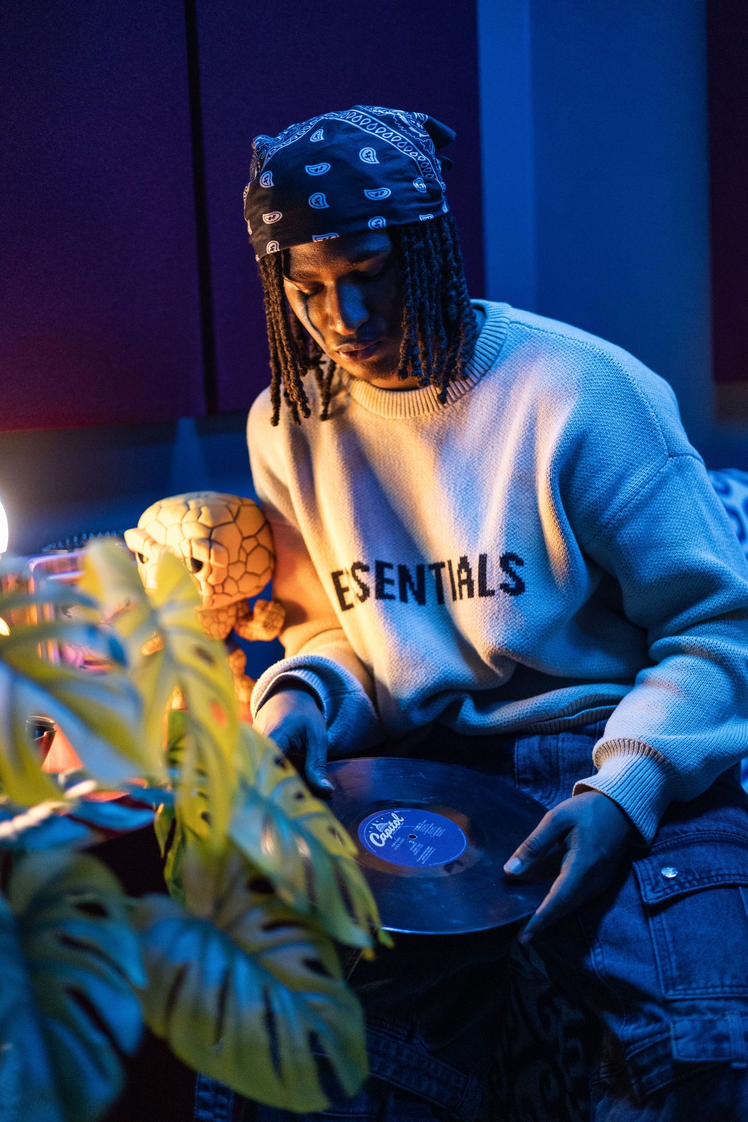 Young man wearing Essentials sweater and bandana holds vinyl record in blue-lit studio with tropical plant.