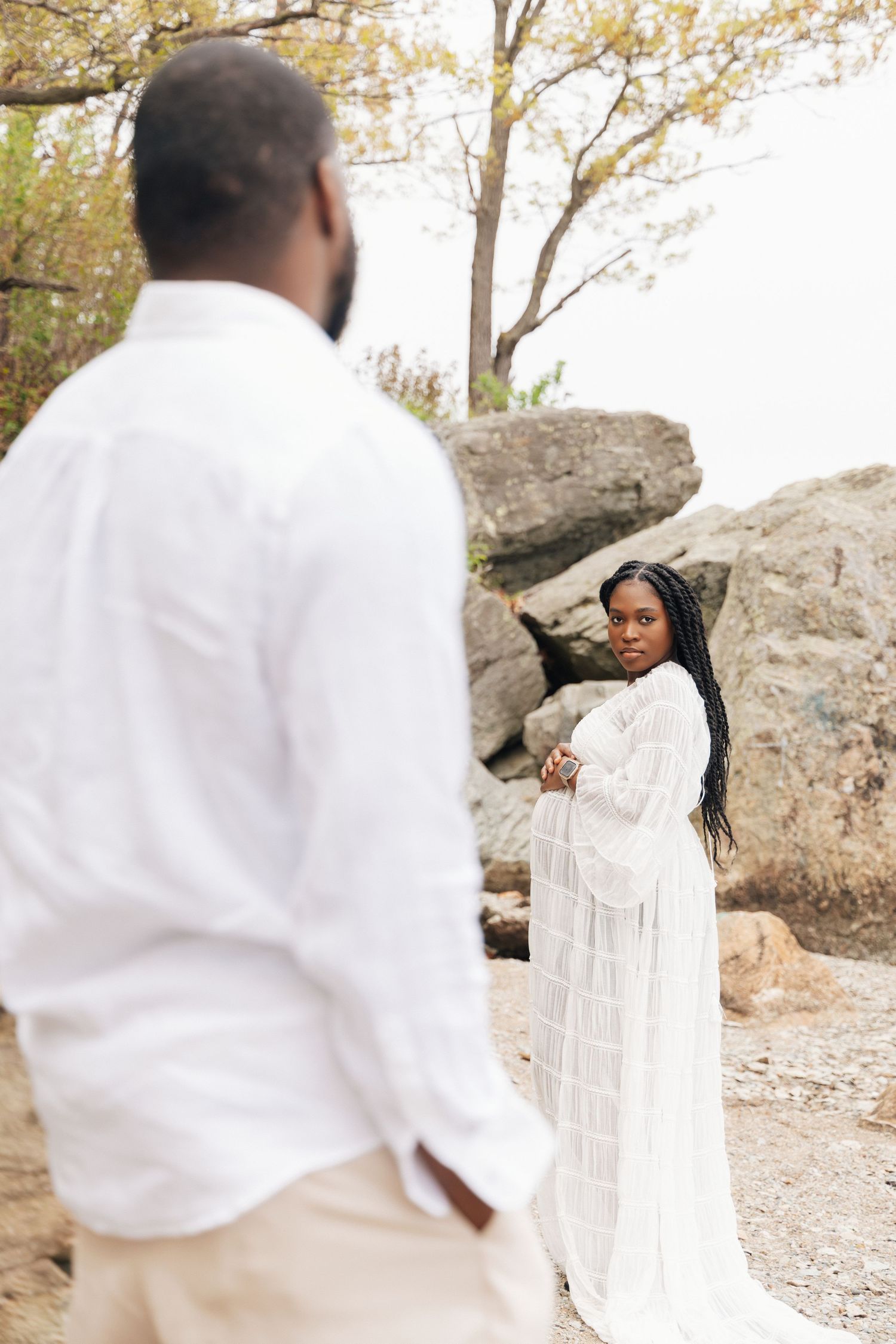 A Black couple in white outfits poses near rocky terrain, woman in lace dress looks back at man in linen shirt.