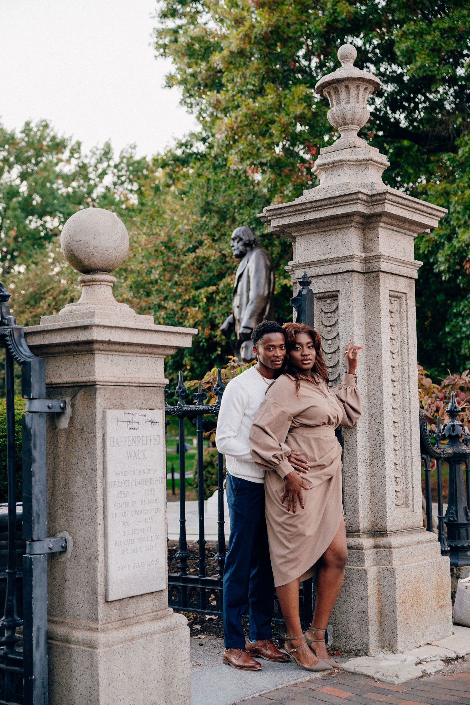 Elegant couple posing at ornate stone gate with bronze statue, surrounded by lush green trees.