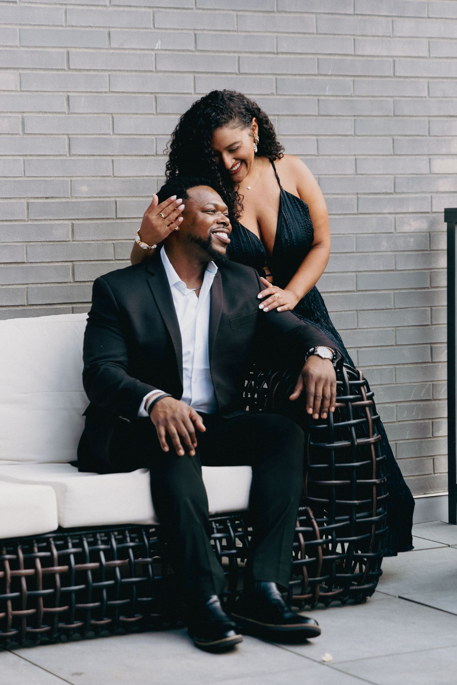 Elegant couple in black formal wear posing together on a wicker sofa against a gray brick wall.