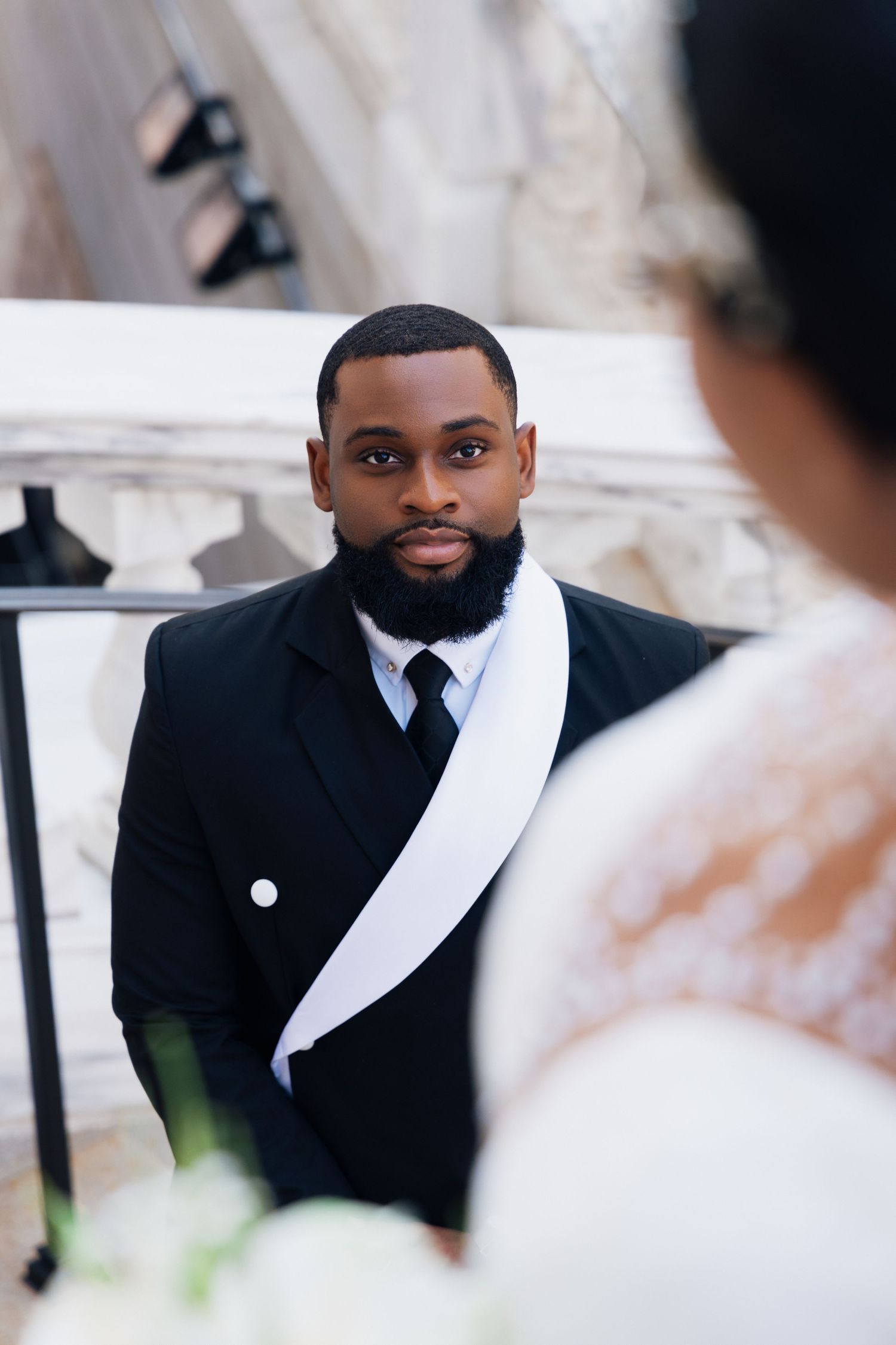 A groom in a black suit with white sash gazes at his bride during an outdoor wedding ceremony.