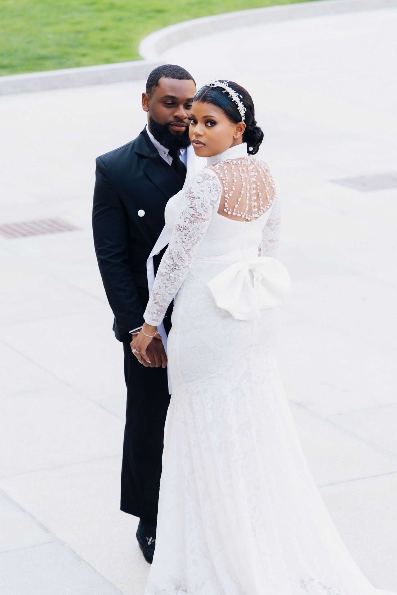Elegant Black couple in wedding attire, bride in white lace gown with pearl back and bow, groom in black suit, posing outdoors.