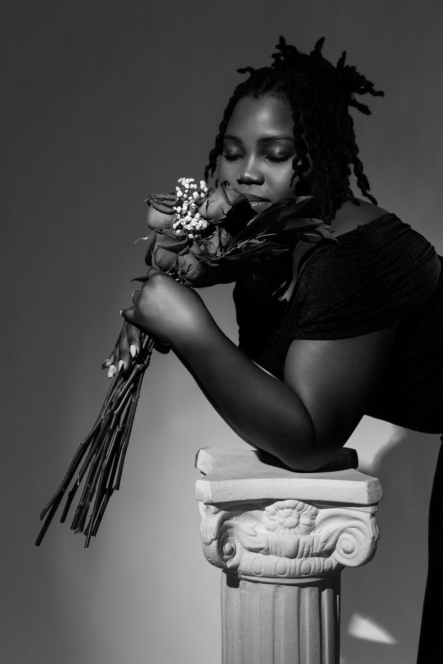Black and white portrait of a woman with dreadlocks leaning on a decorative column holding flowers.