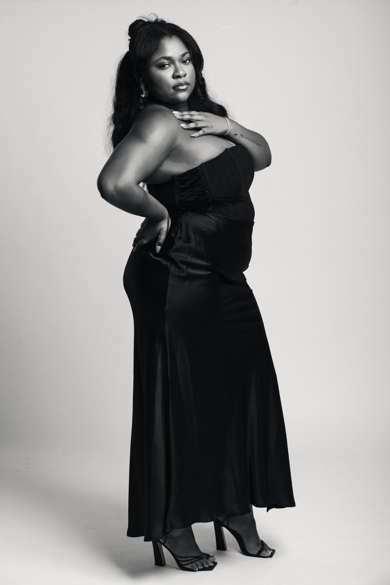 Black and white photo of a woman in an elegant strapless black gown and heels posing confidently in a studio.