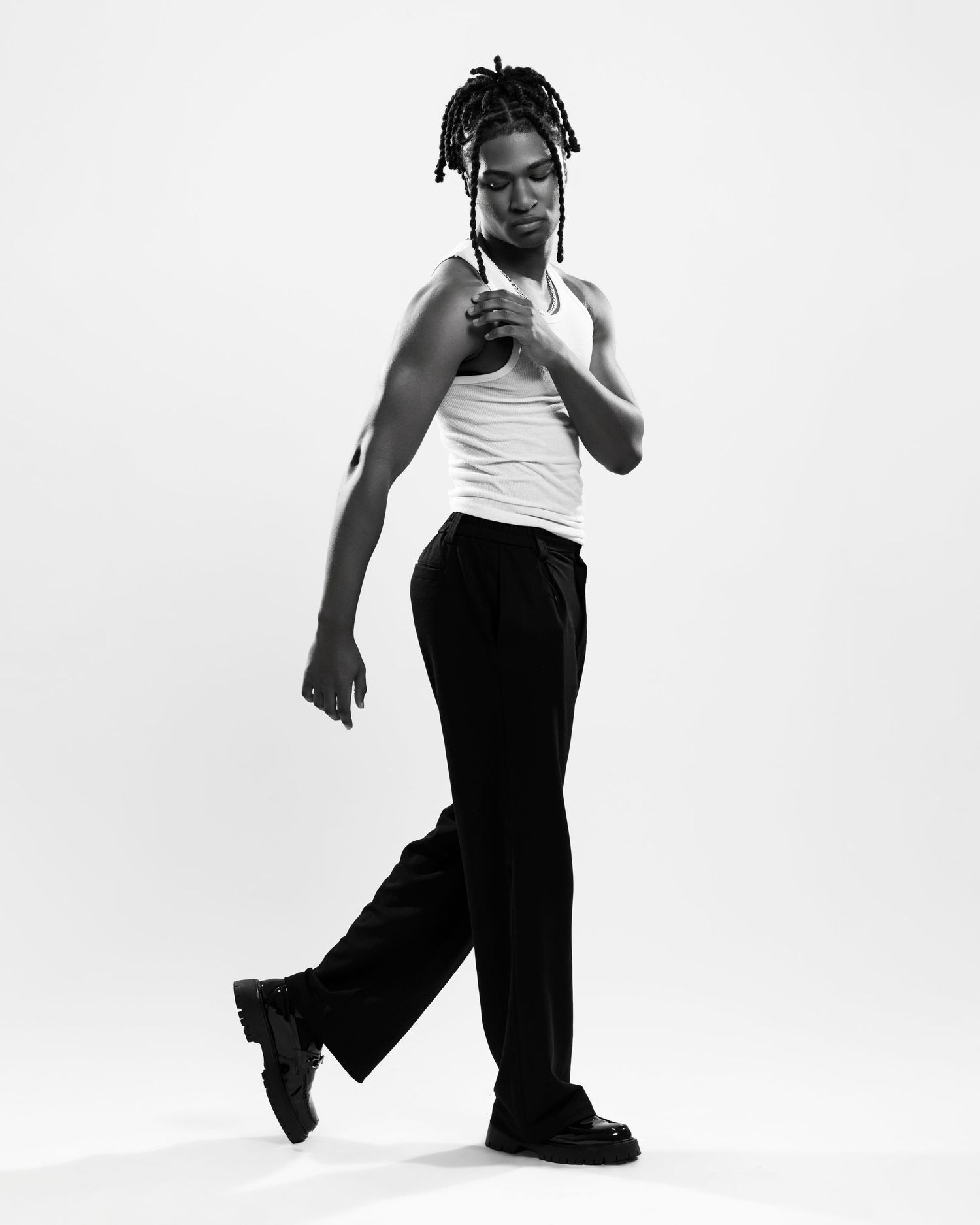 Black and white photo of a young man with locs wearing a white tank top and black trousers, posing dynamically.