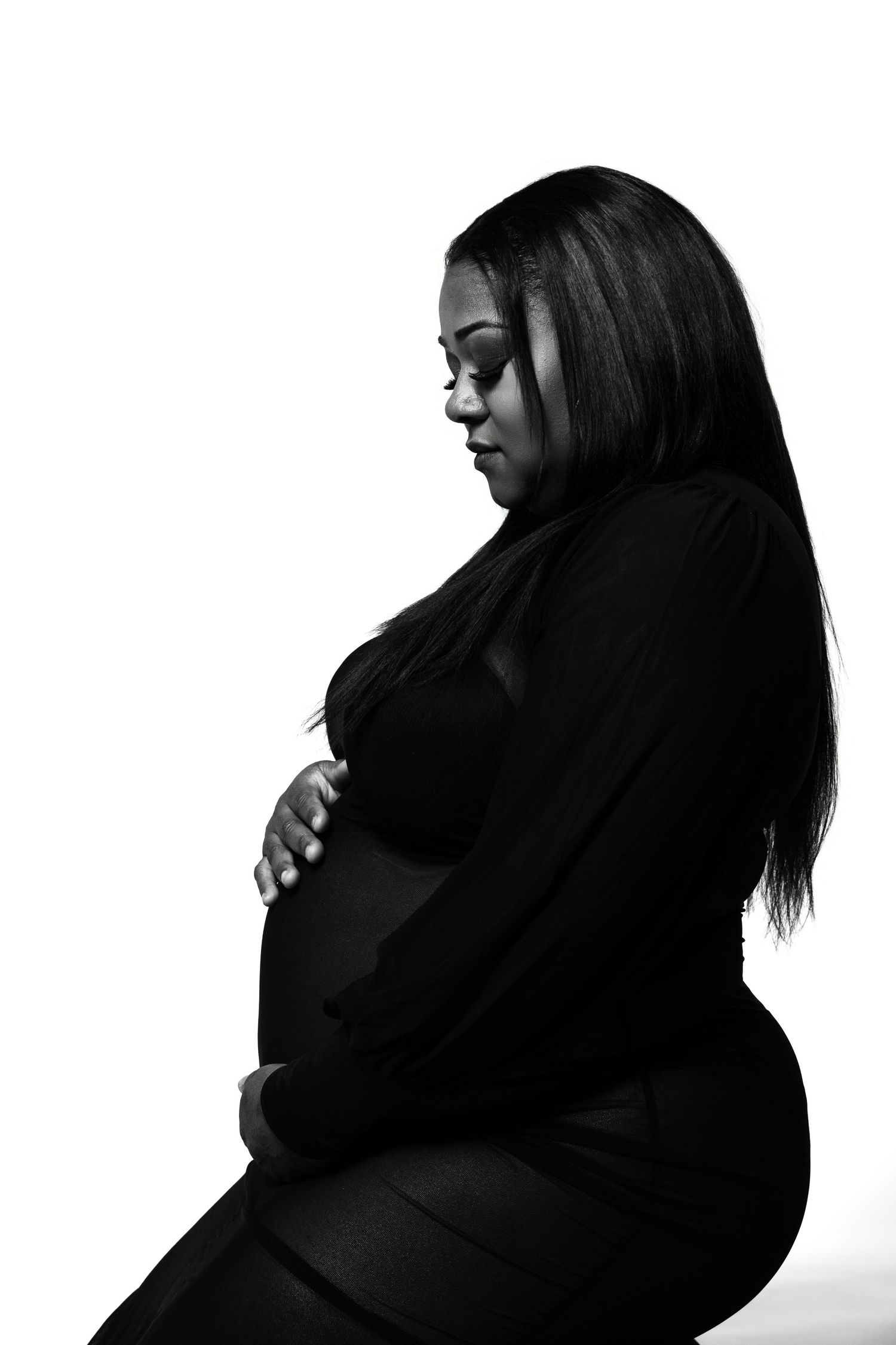 Black and white maternity photo of a pregnant woman in black outfit cradling her baby bump against white background.