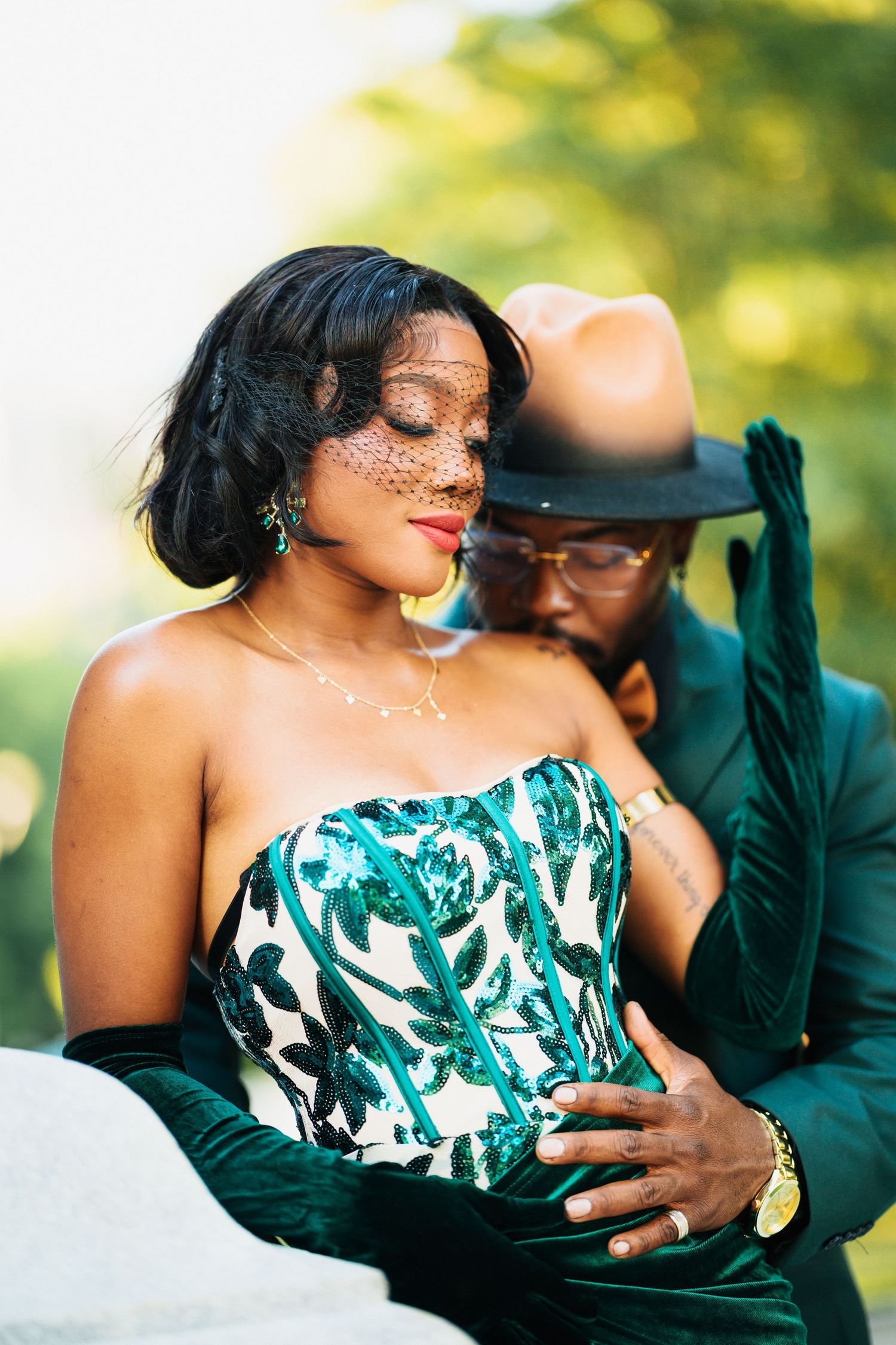 Elegant couple in matching green outfits, woman in sequined corset with veil, man in hat embracing her outdoors.