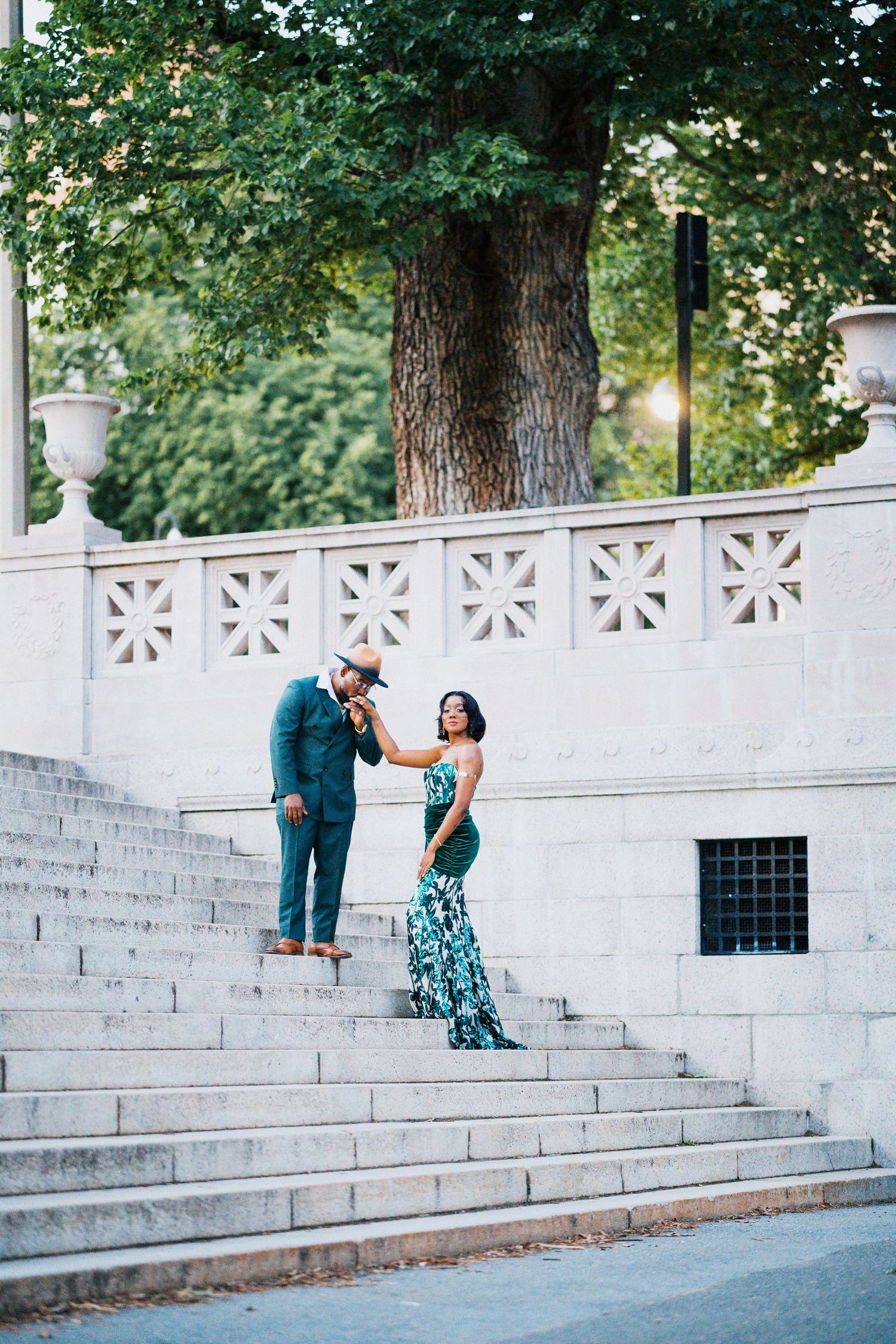 Couple in elegant green attire share a romantic moment on white marble steps outdoors.
