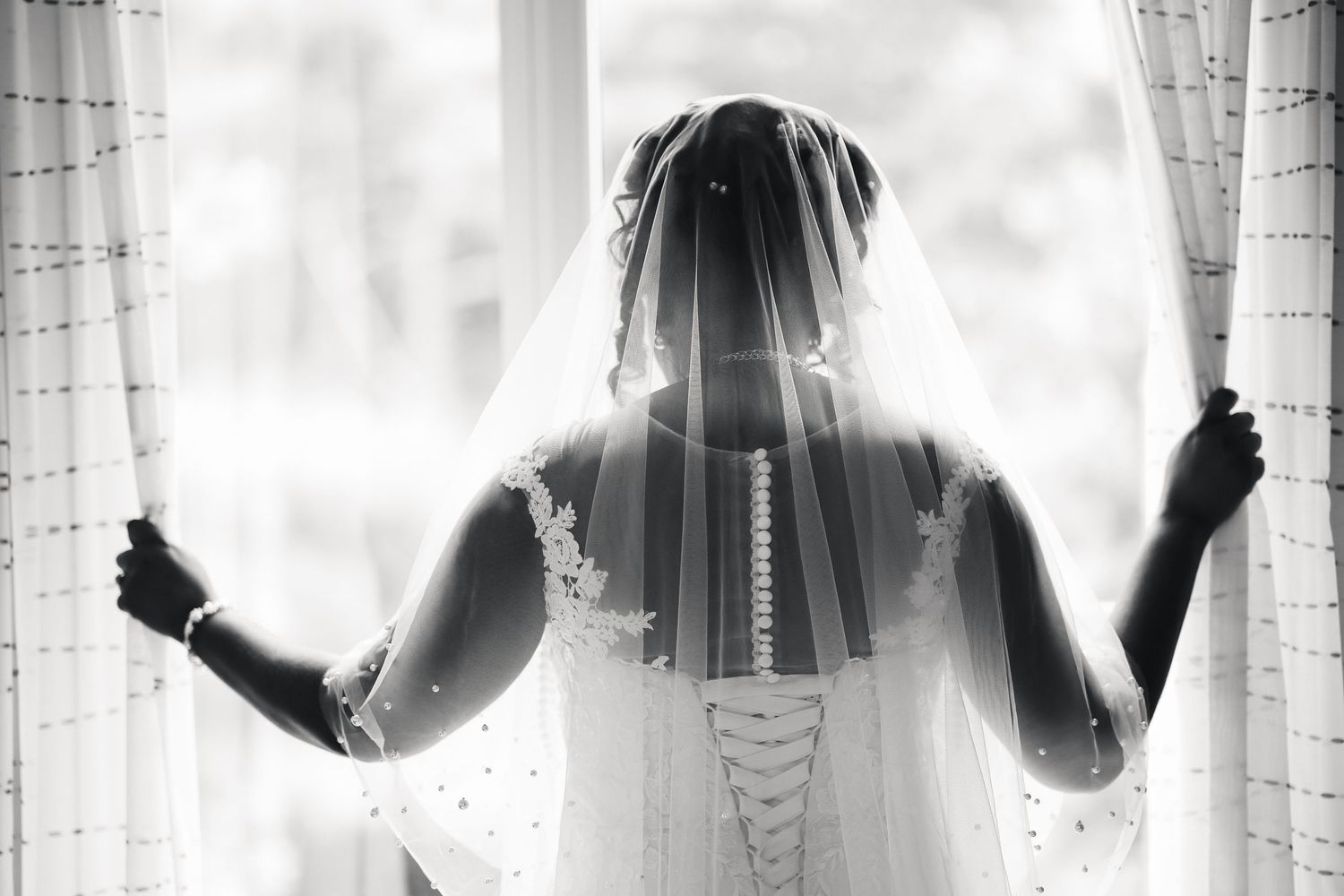 Black and white photo of a bride in lace wedding dress and veil standing at a sunlit window.