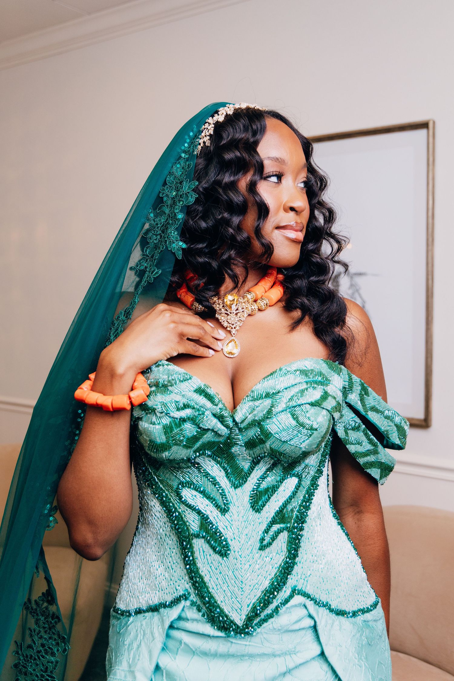 Bride in teal beaded off-shoulder gown with lace veil and orange coral jewelry looking sideways.
