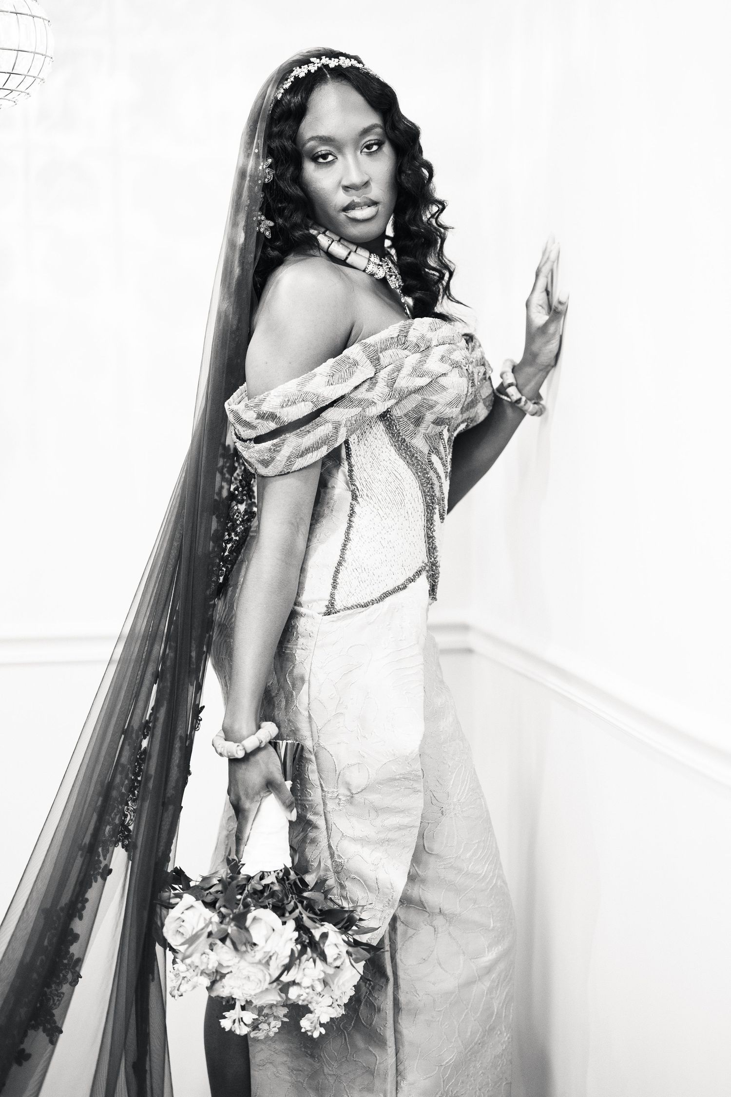 Black and white portrait of bride in off-shoulder gown holding bouquet by staircase.