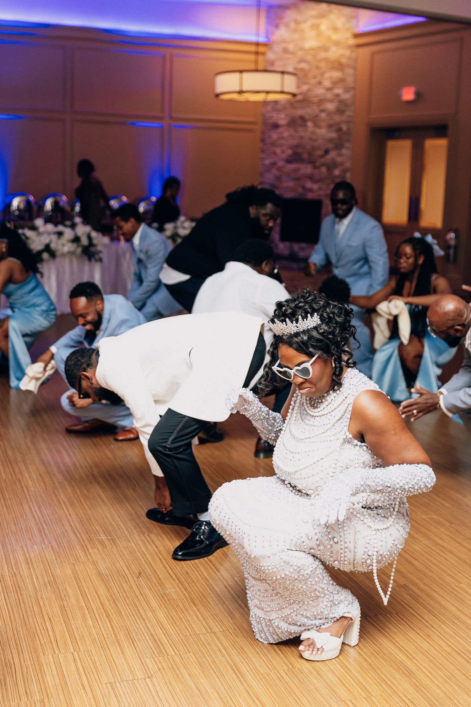 Bride and groom joyfully dancing at wedding reception, bride wearing beaded gown and fun sunglasses on dance floor.