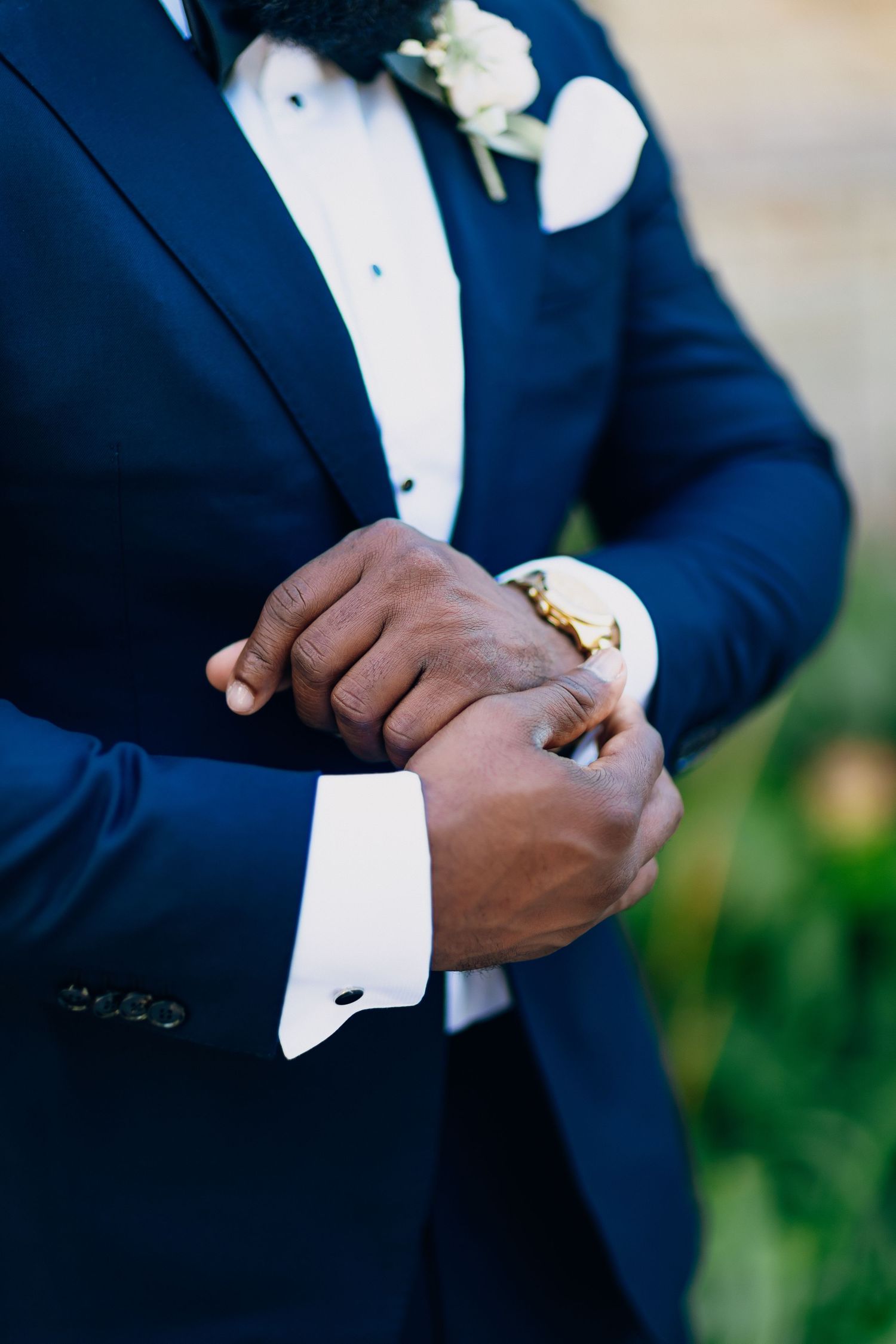 Groom in navy tuxedo adjusts gold cufflinks on white dress shirt with boutonniere detail.