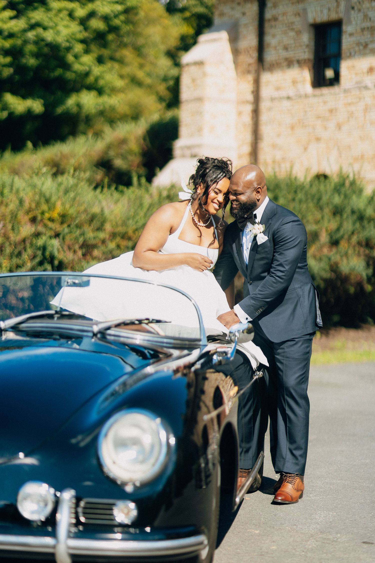 A bride and groom laugh together while leaning over a classic black convertible outdoors.