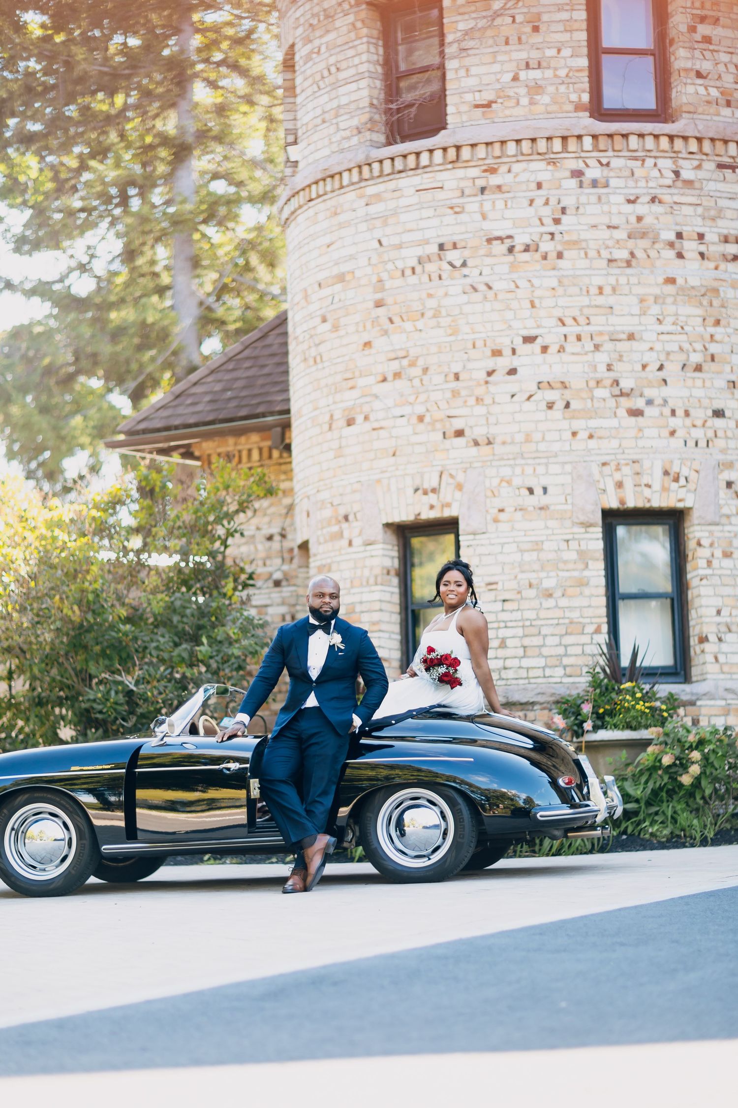 Bride and groom pose separately beside a vintage black convertible in front of a brick tower.