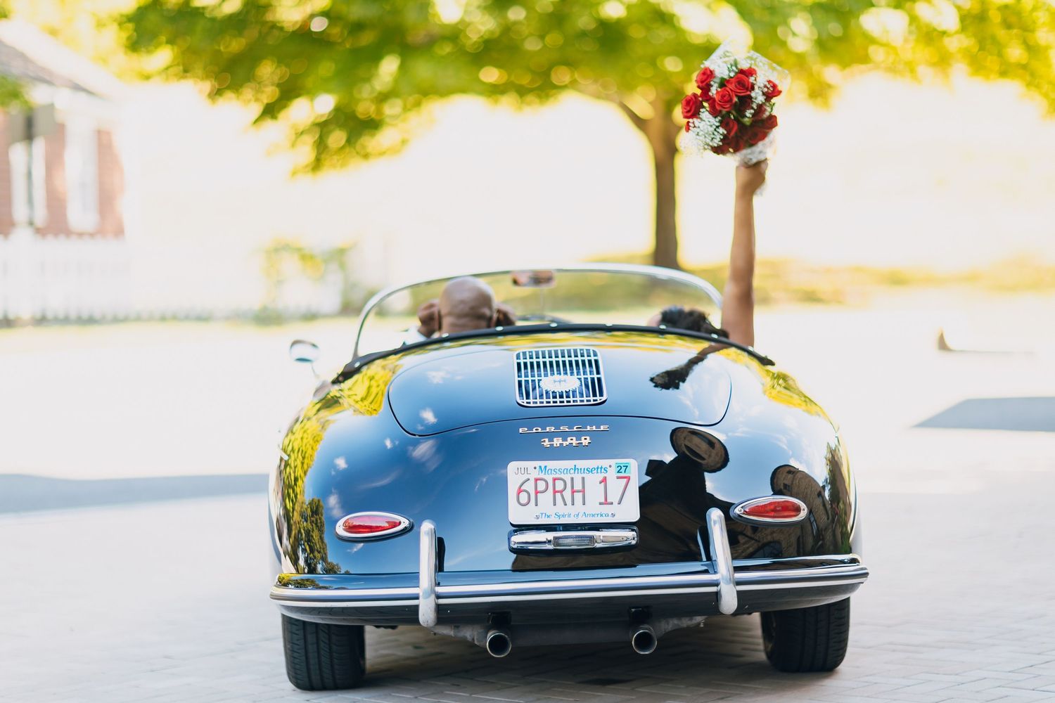 Rear view of newlyweds driving away in a classic black Porsche 356 convertible with red bouquet.