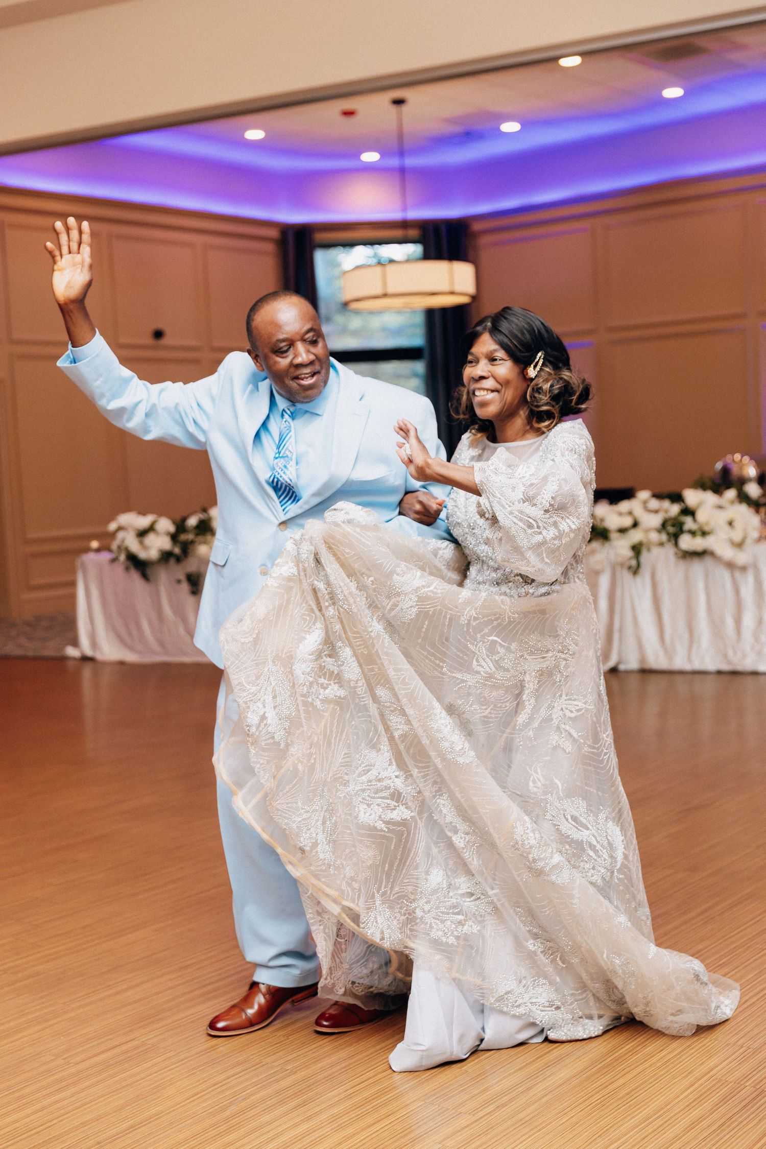 Joyful bride and groom dancing energetically at wedding reception in elegant ballroom with purple uplighting.